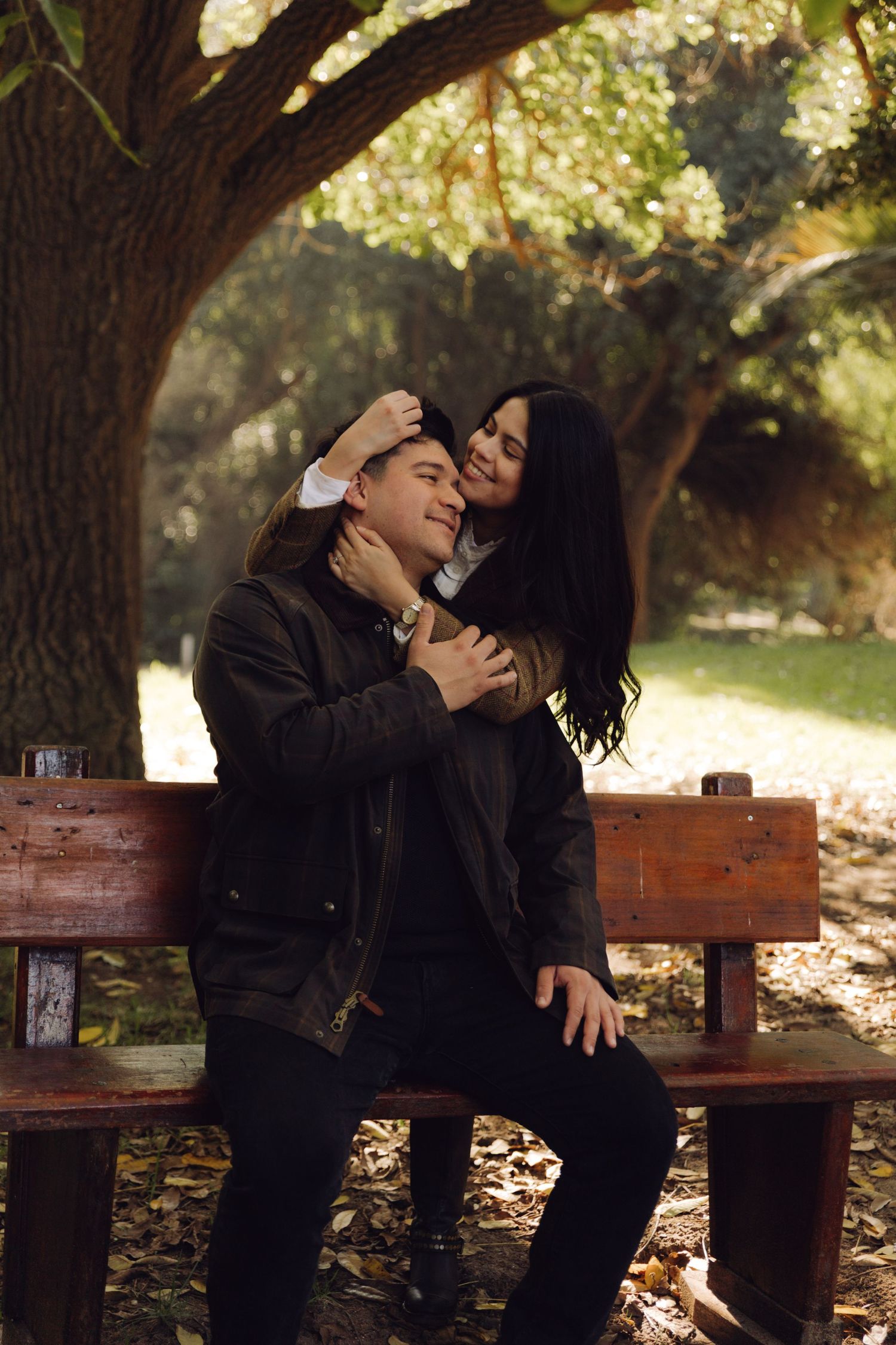 Couple sharing a tender moment on a wooden park bench, woman embracing man from behind under autumn trees.