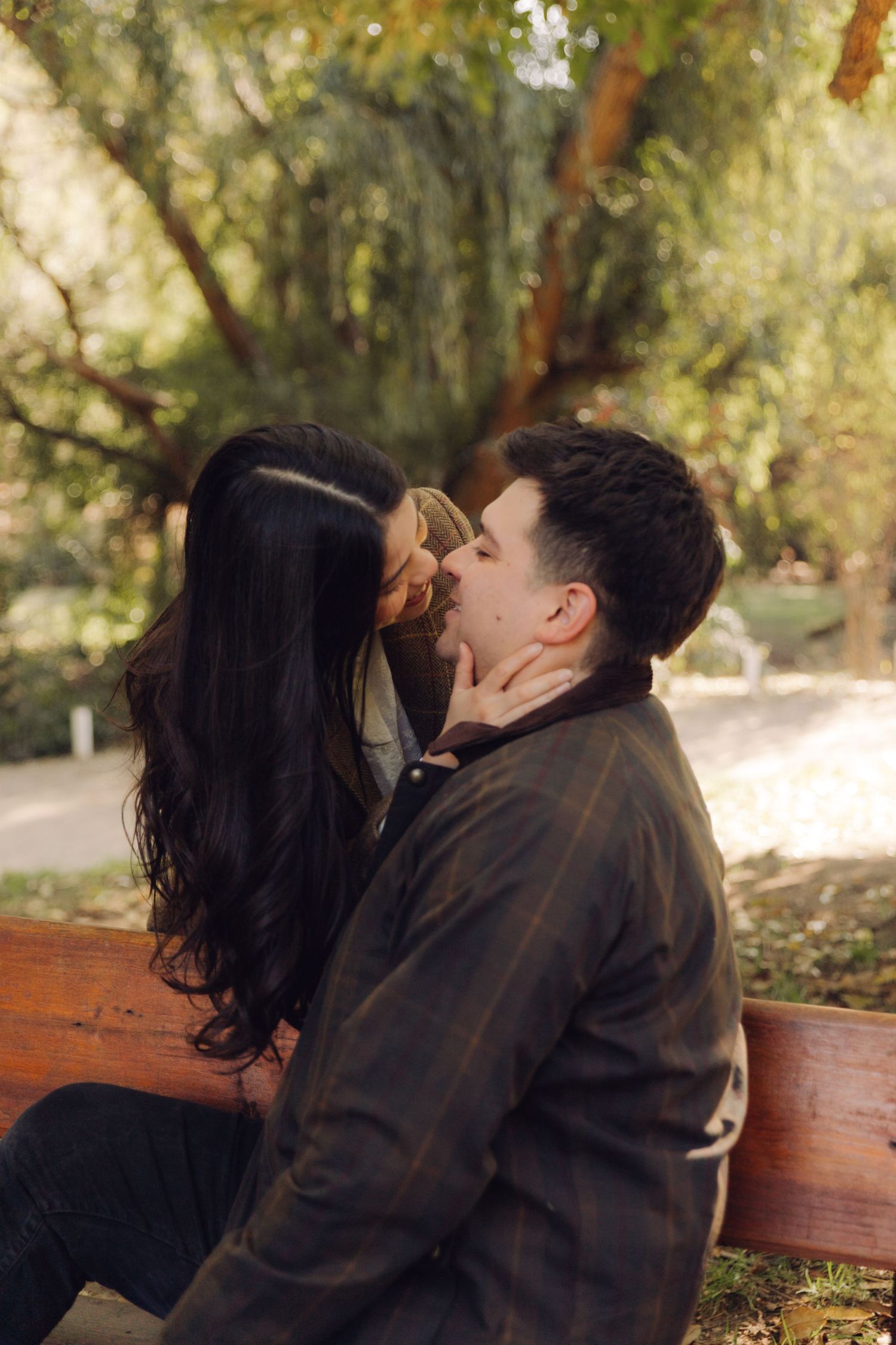 Romantic couple sharing a kiss on a wooden bench in a lush green park setting.