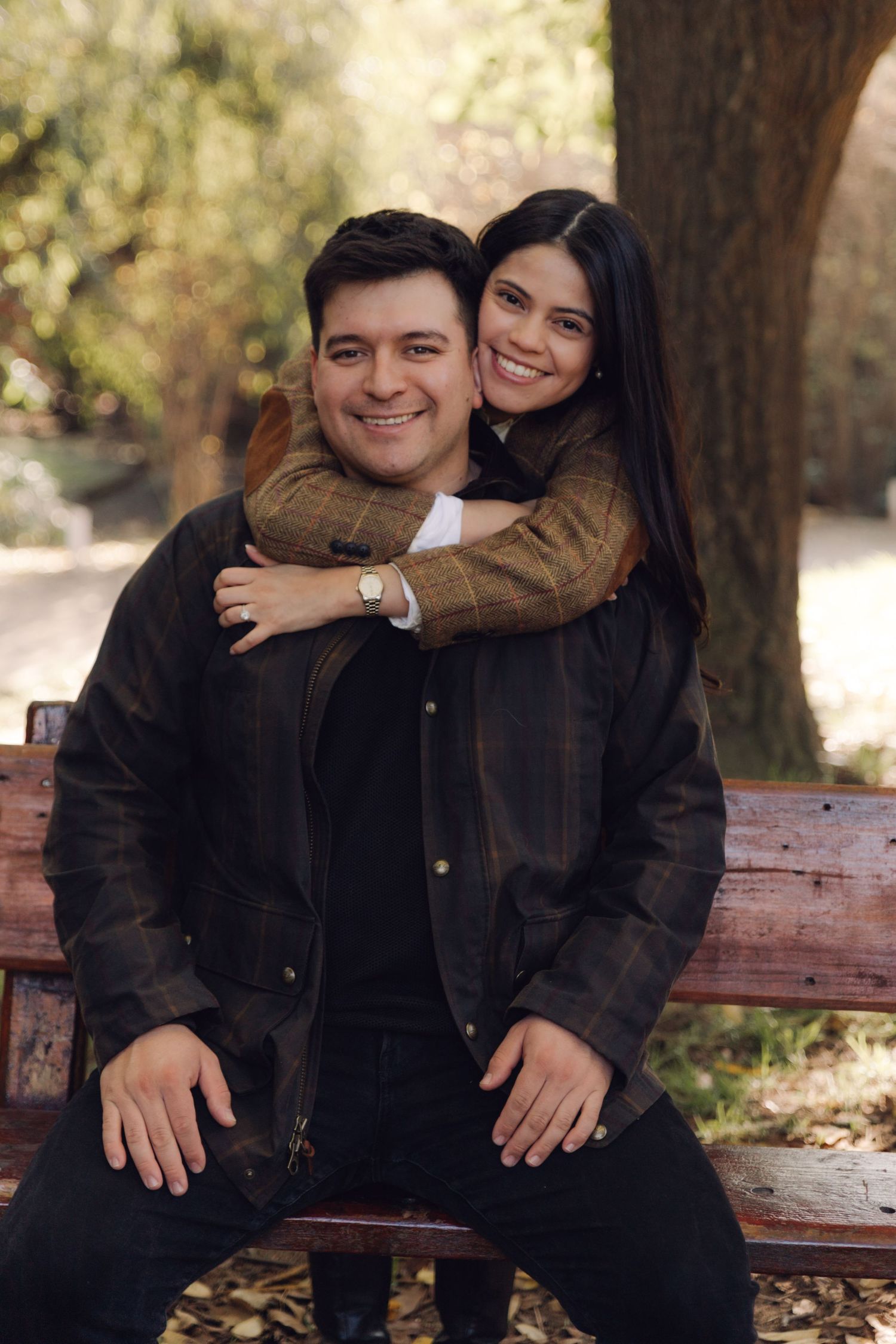Smiling couple posing on a wooden park bench, woman hugging man from behind among autumn trees.