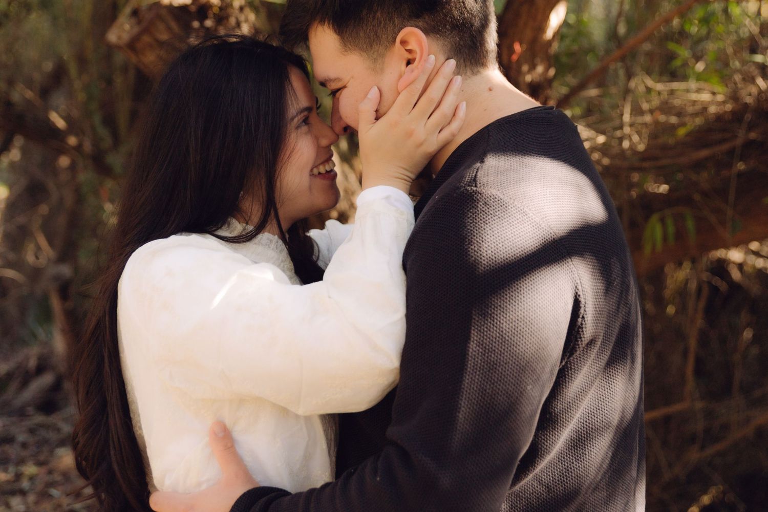 Couple sharing a tender romantic moment outdoors, woman holding mans face gently in dappled sunlight.