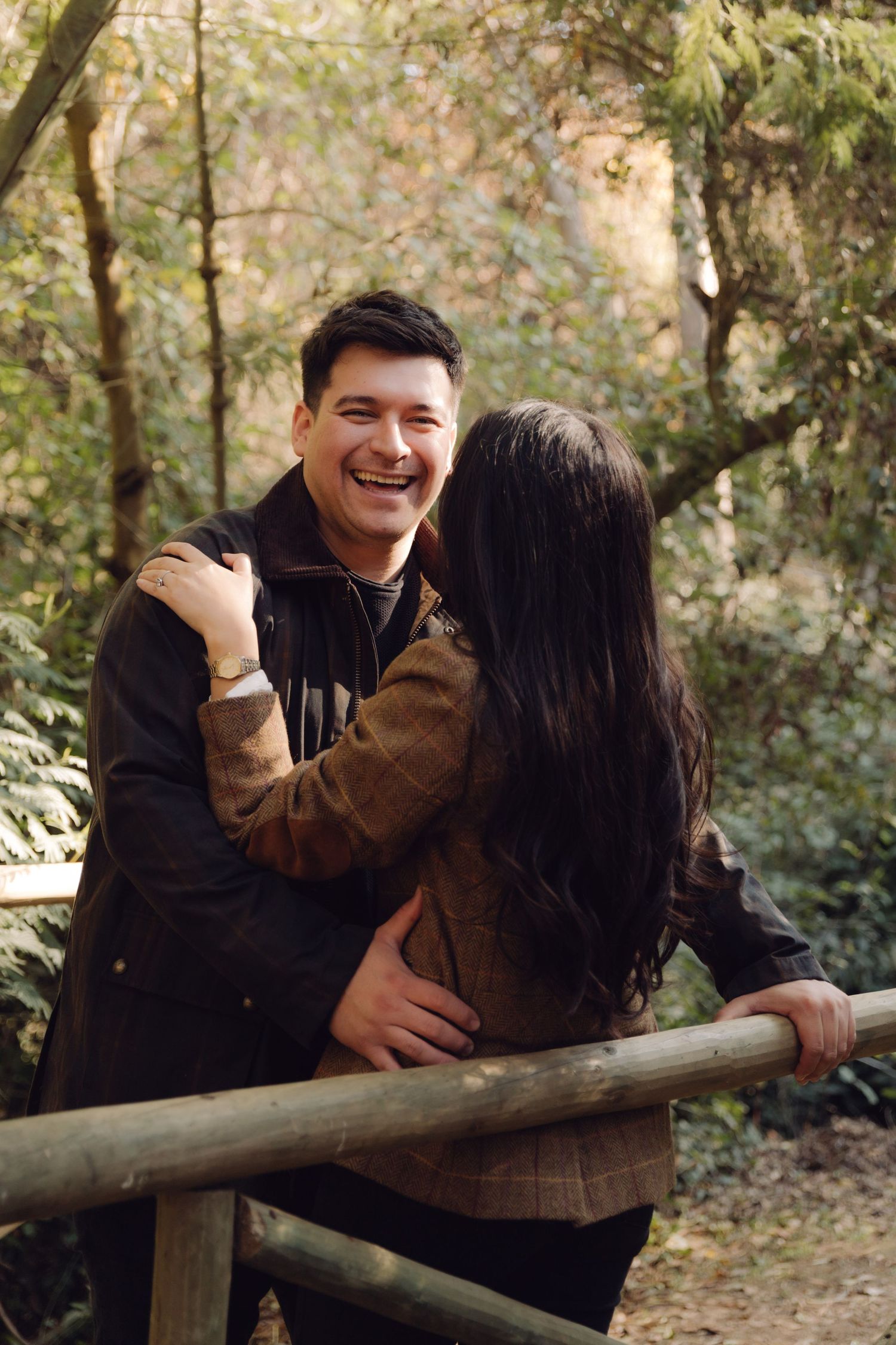 Laughing man embraces woman on wooden bridge in autumn forest park during romantic outdoor couples photoshoot.