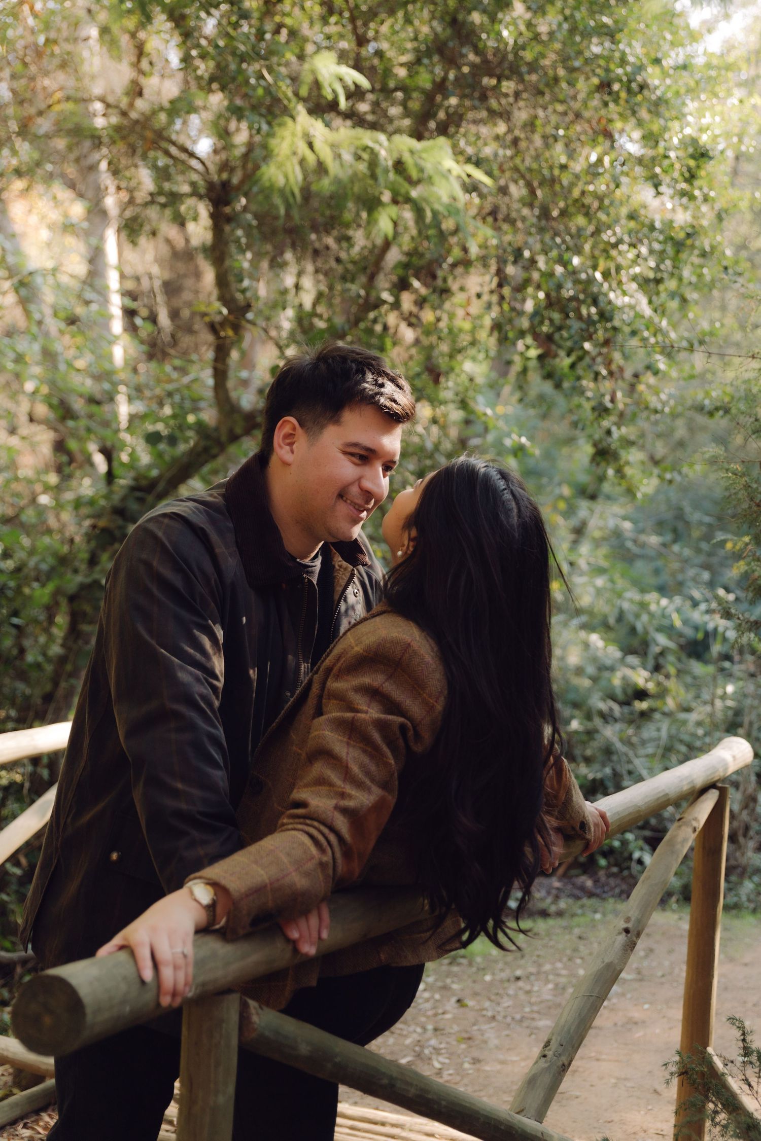 Romantic couple almost kissing on rustic wooden bridge surrounded by lush green trees in a park.