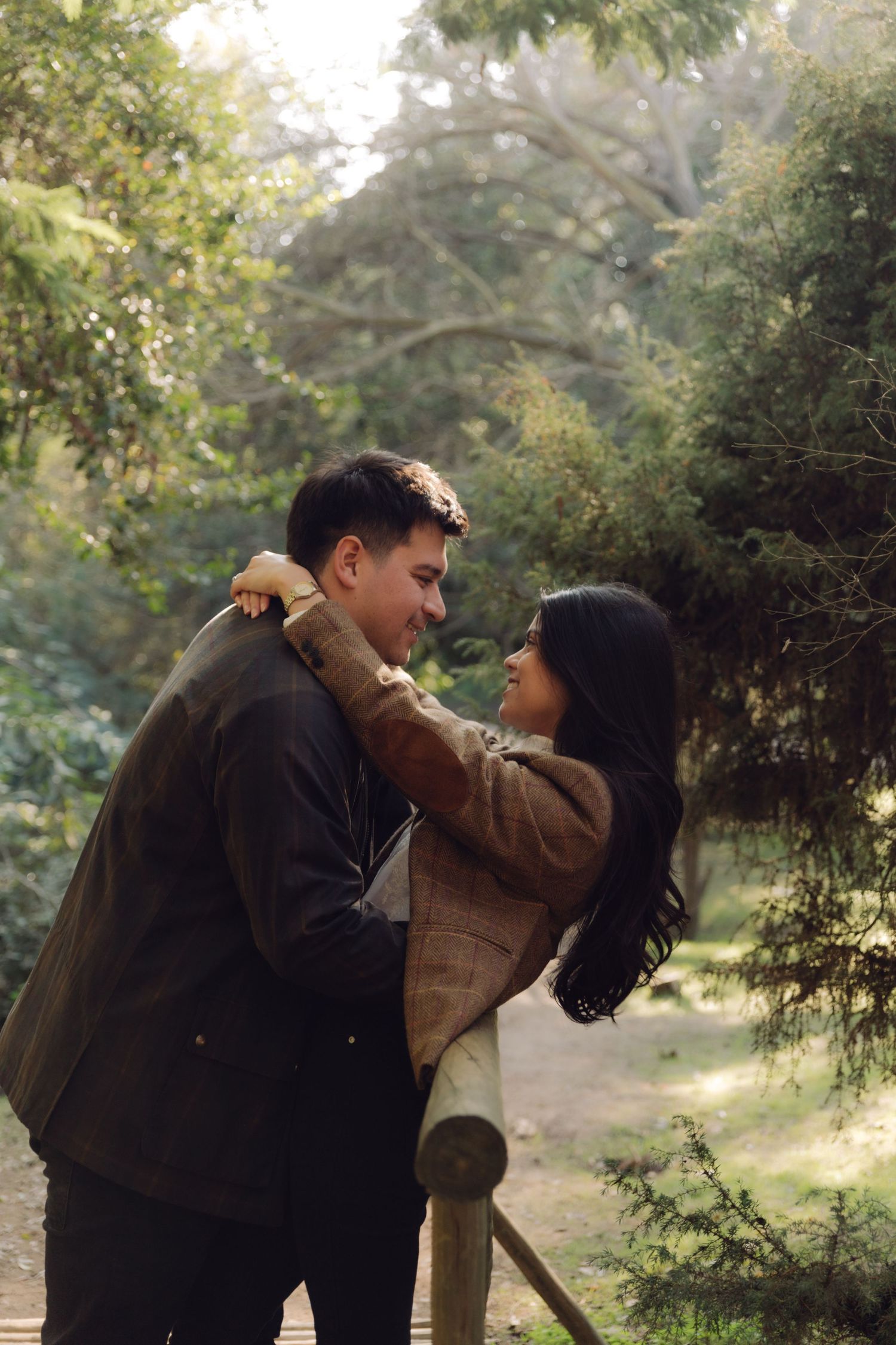 Happy couple embracing and smiling at each other on a wooden bridge in a sunlit green forest.