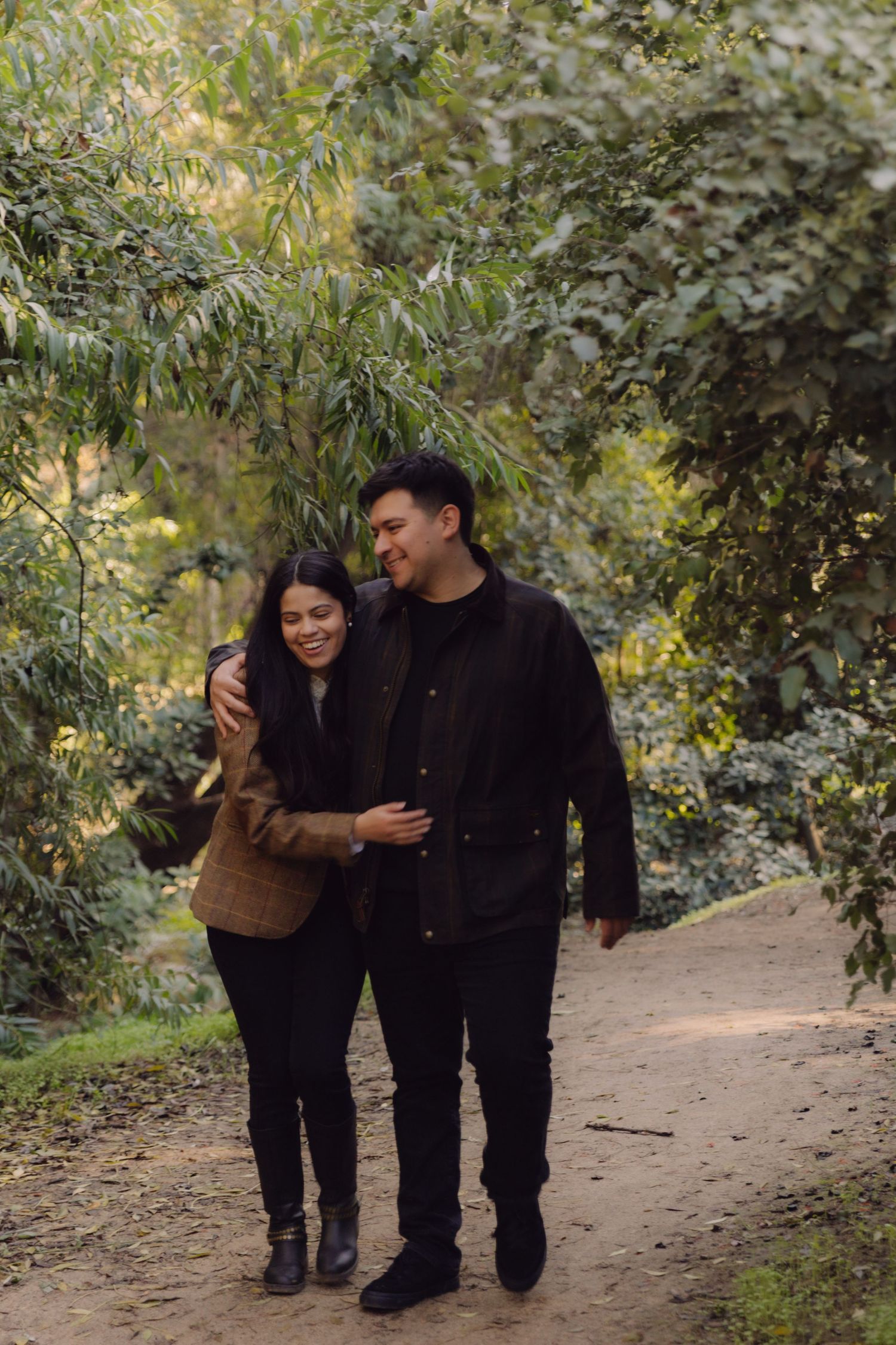 Smiling couple walking arm in arm along a dirt path surrounded by lush green trees in a park.