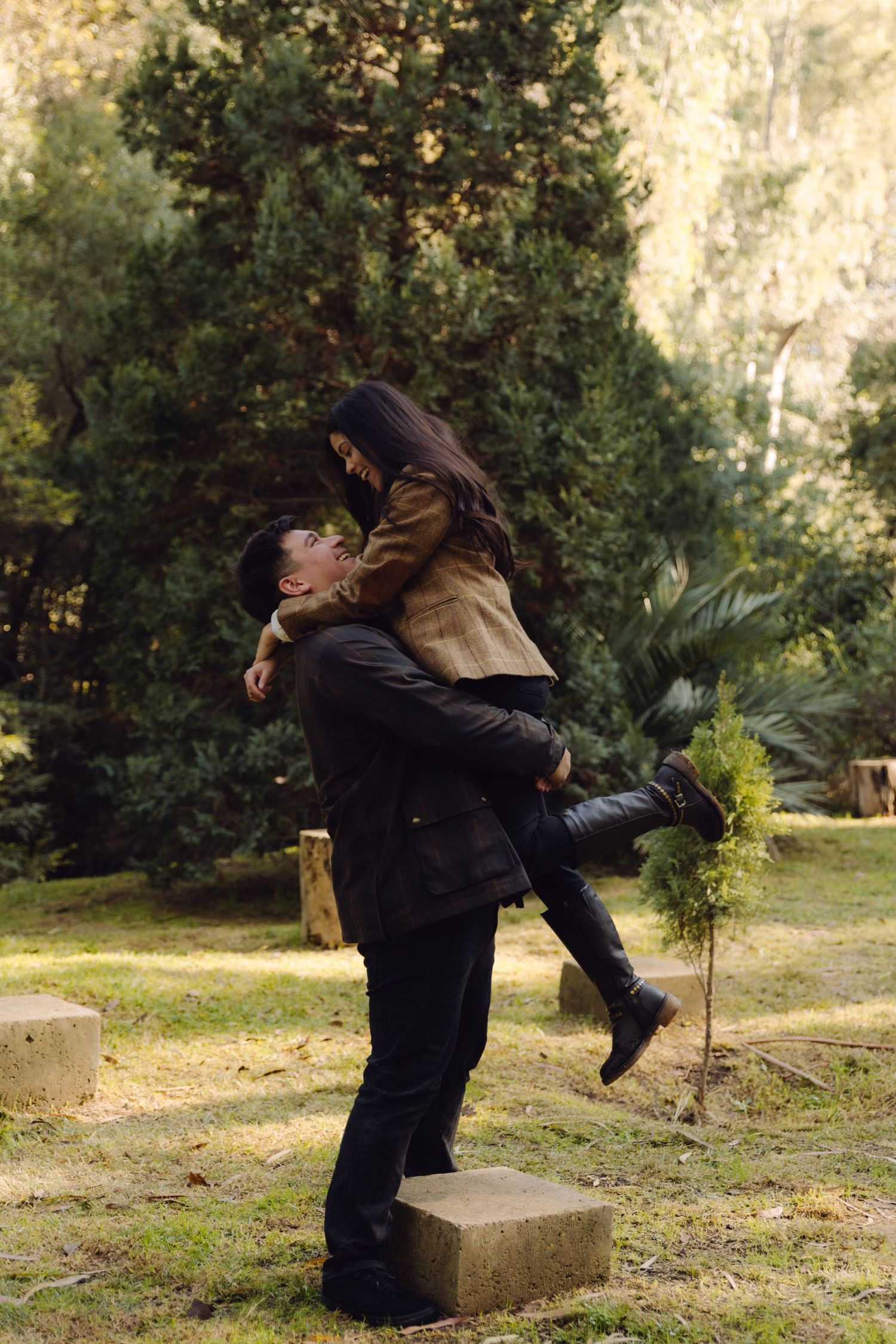 Man lifting woman in his arms while standing on stone block in a sunny green park setting.
