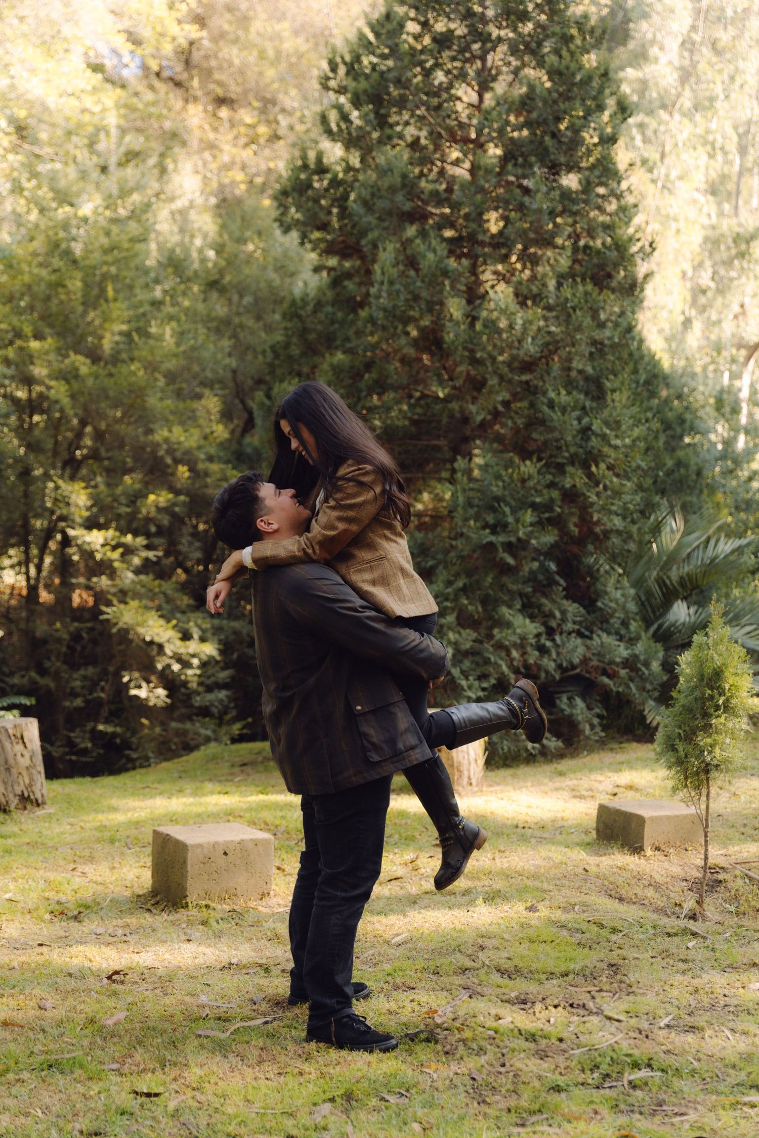 Man lifting smiling woman in his arms outdoors in a sunny green park with tall evergreen trees.