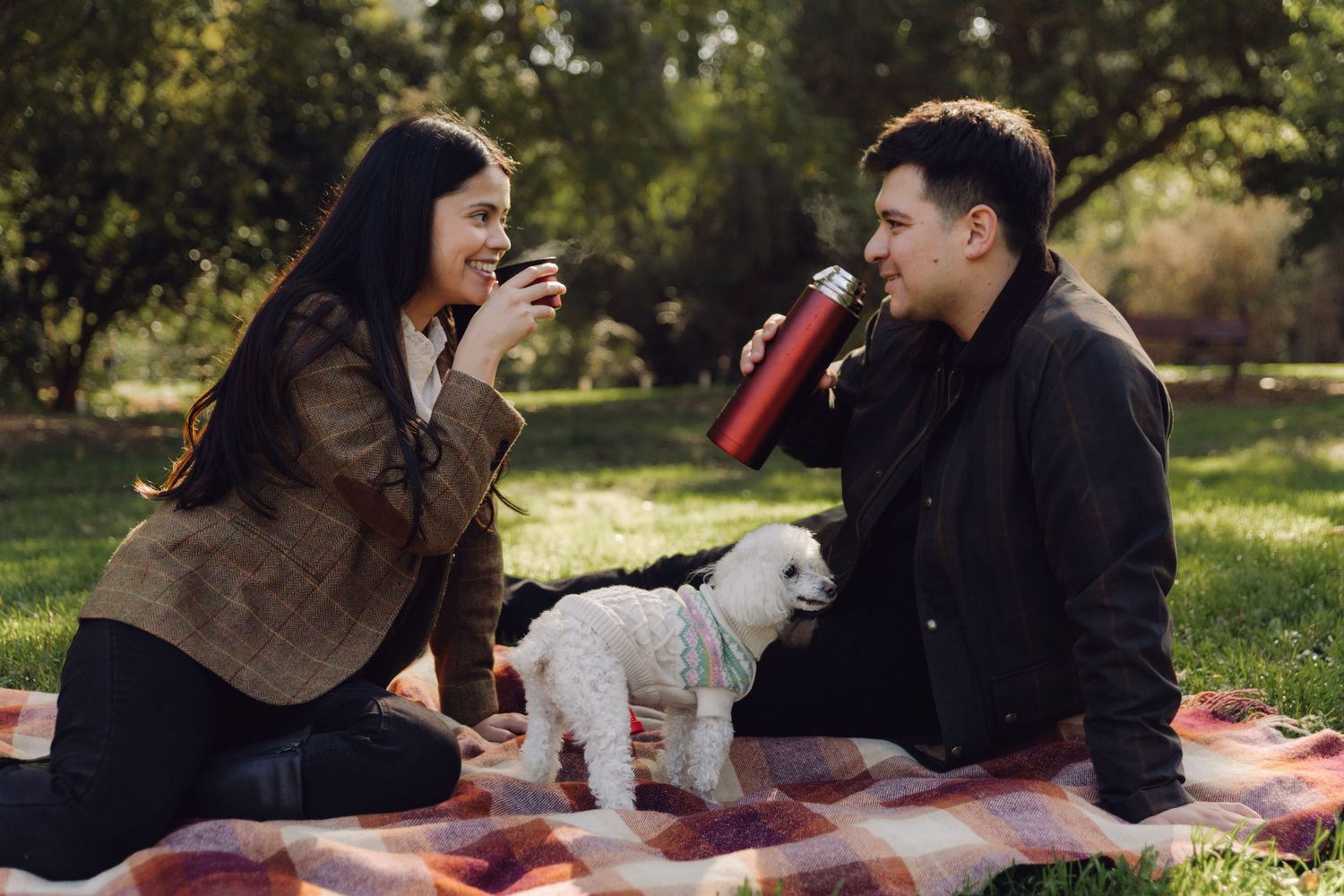 A couple enjoying a picnic on a plaid blanket with a small white dog in the park.