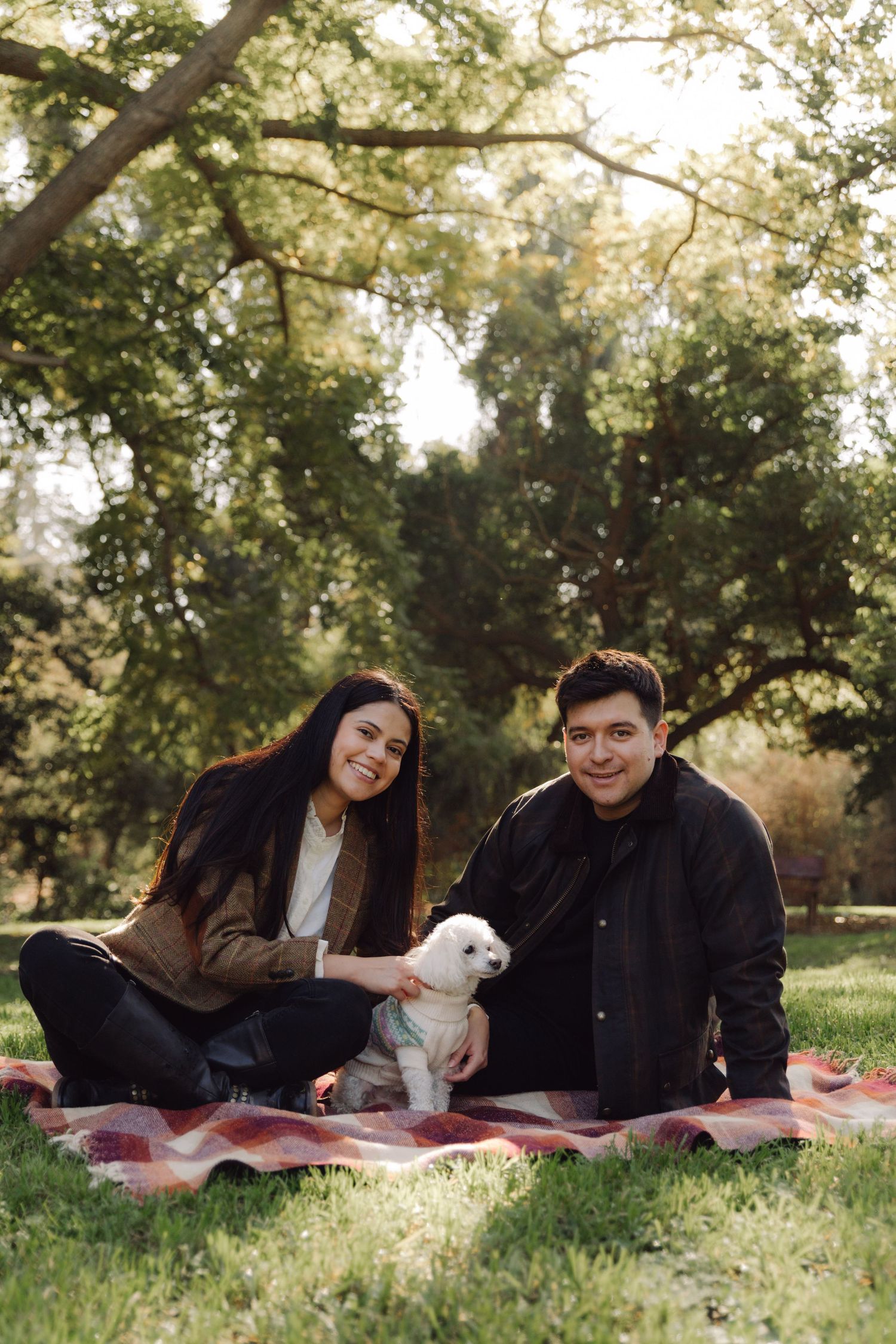 A smiling couple sitting on a plaid blanket in a park with their white fluffy dog.