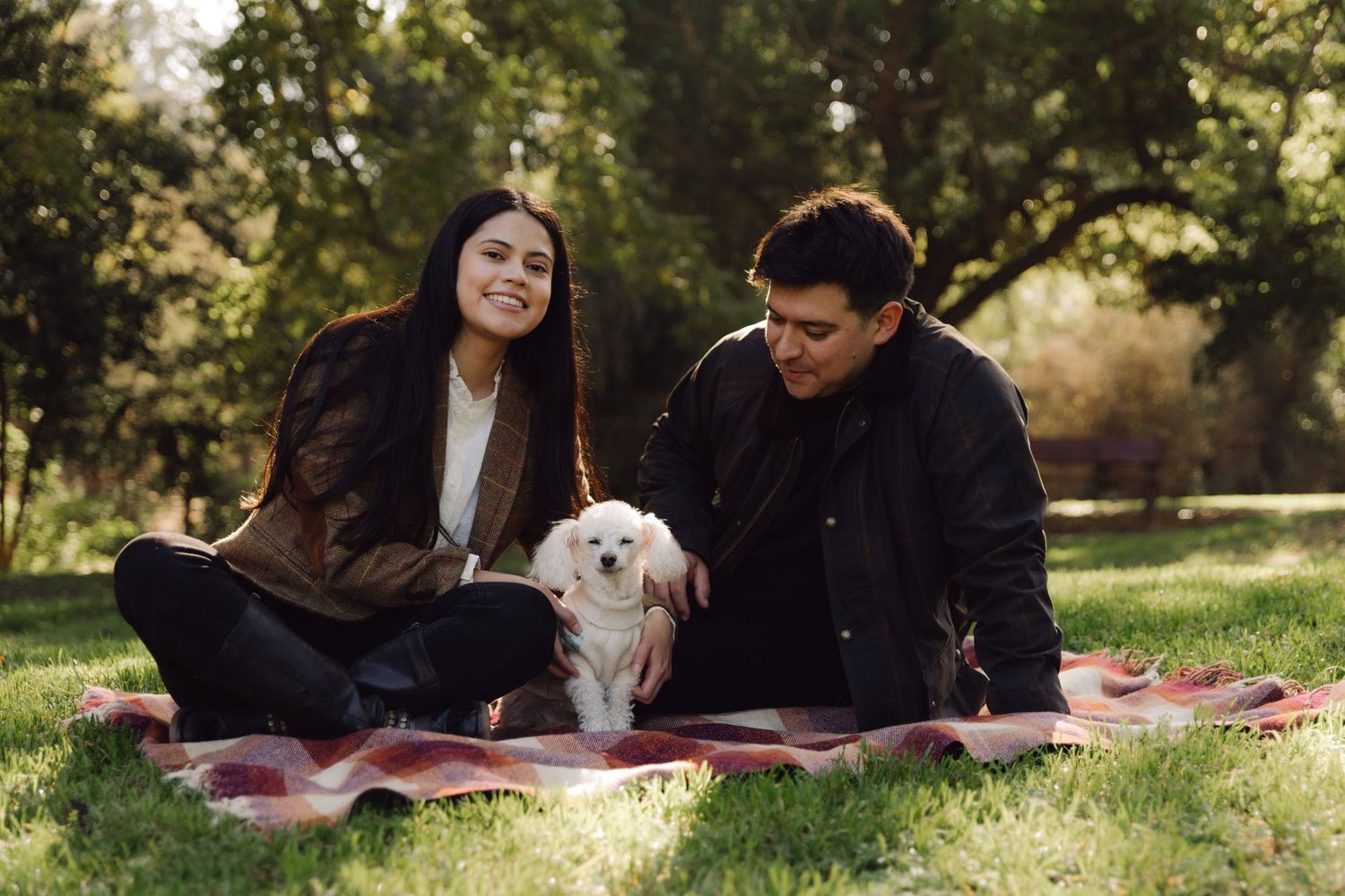 A couple sitting on a plaid blanket in a sunny park with their small white dog.