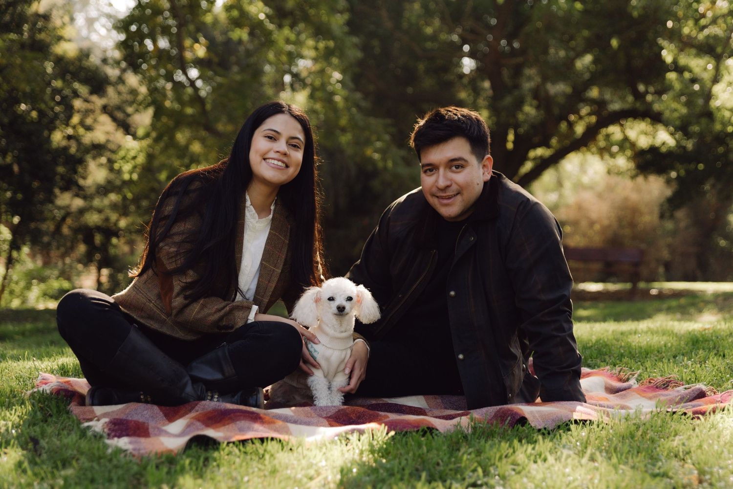 A couple posing on a plaid picnic blanket in a park with their small white dog.