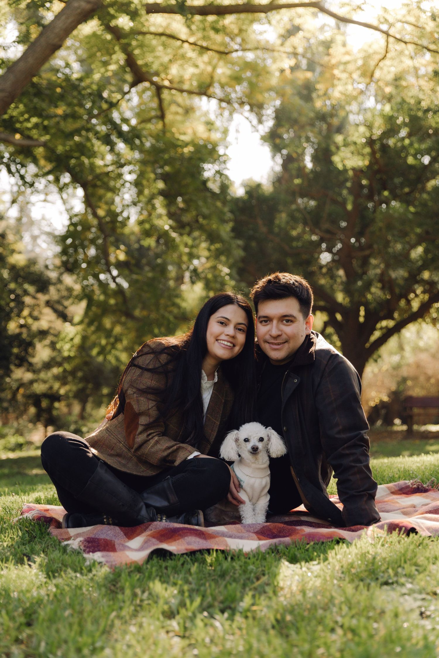 A smiling couple sits on a plaid blanket in a park with their white Bichon Frise dog on a sunny day.