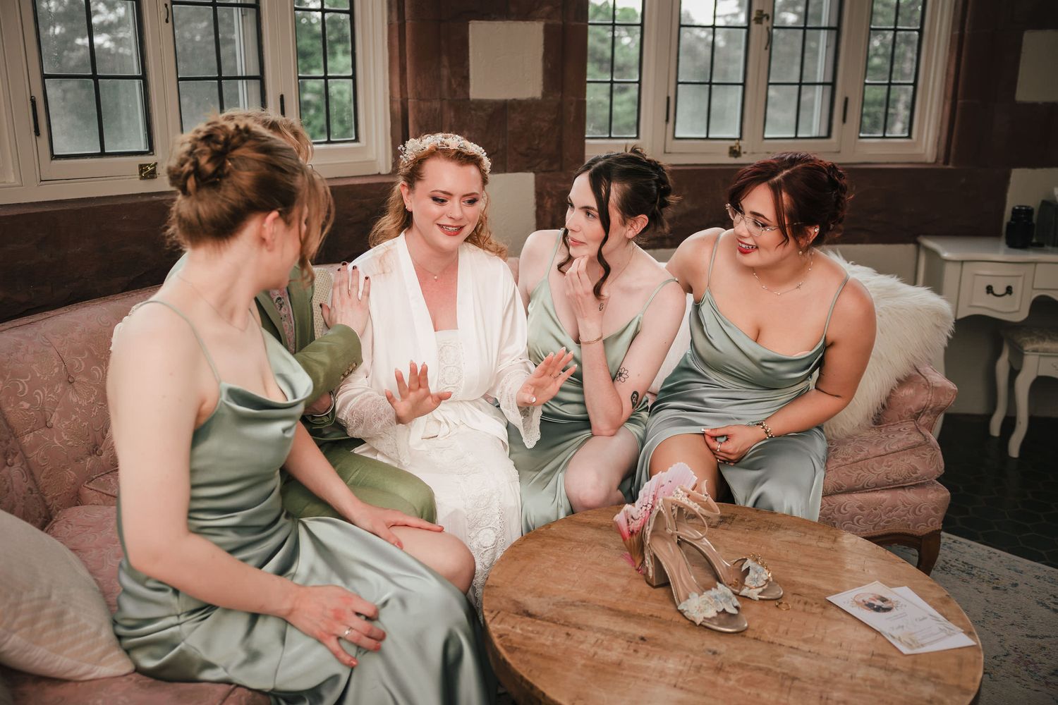 Bride in white robe sits with three bridesmaids in sage green gowns, sharing a joyful moment before the wedding.