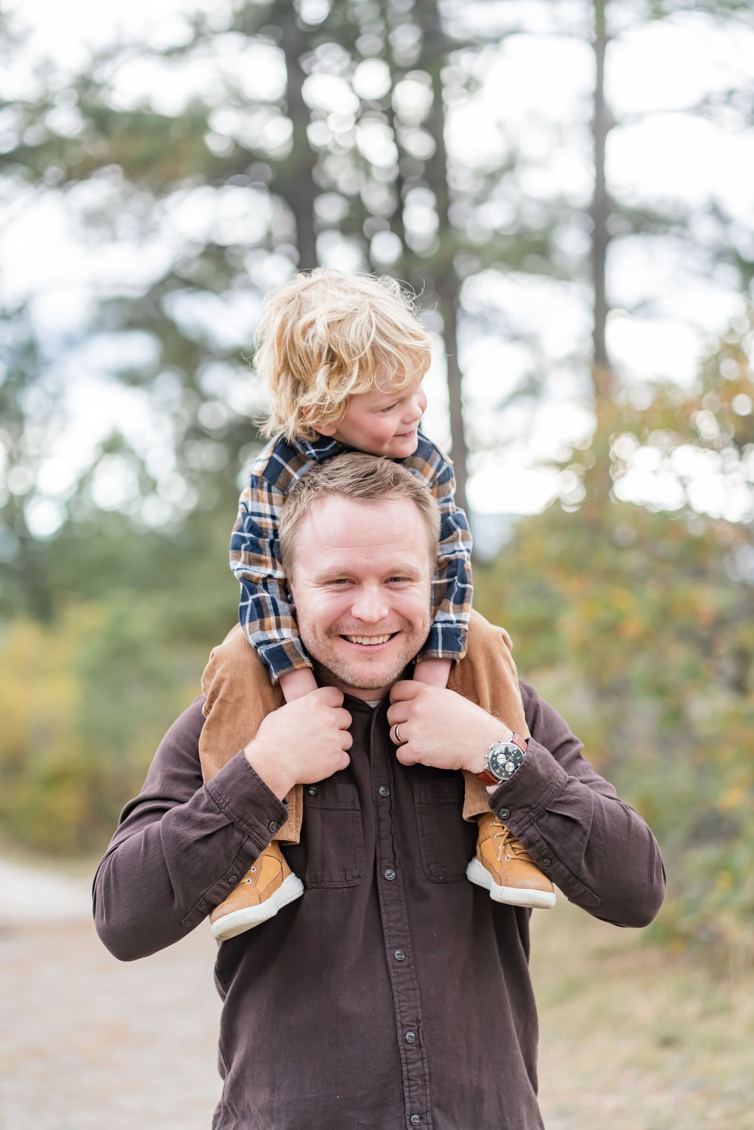 A smiling man carries a young blond boy on his shoulders outdoors in a forest setting.