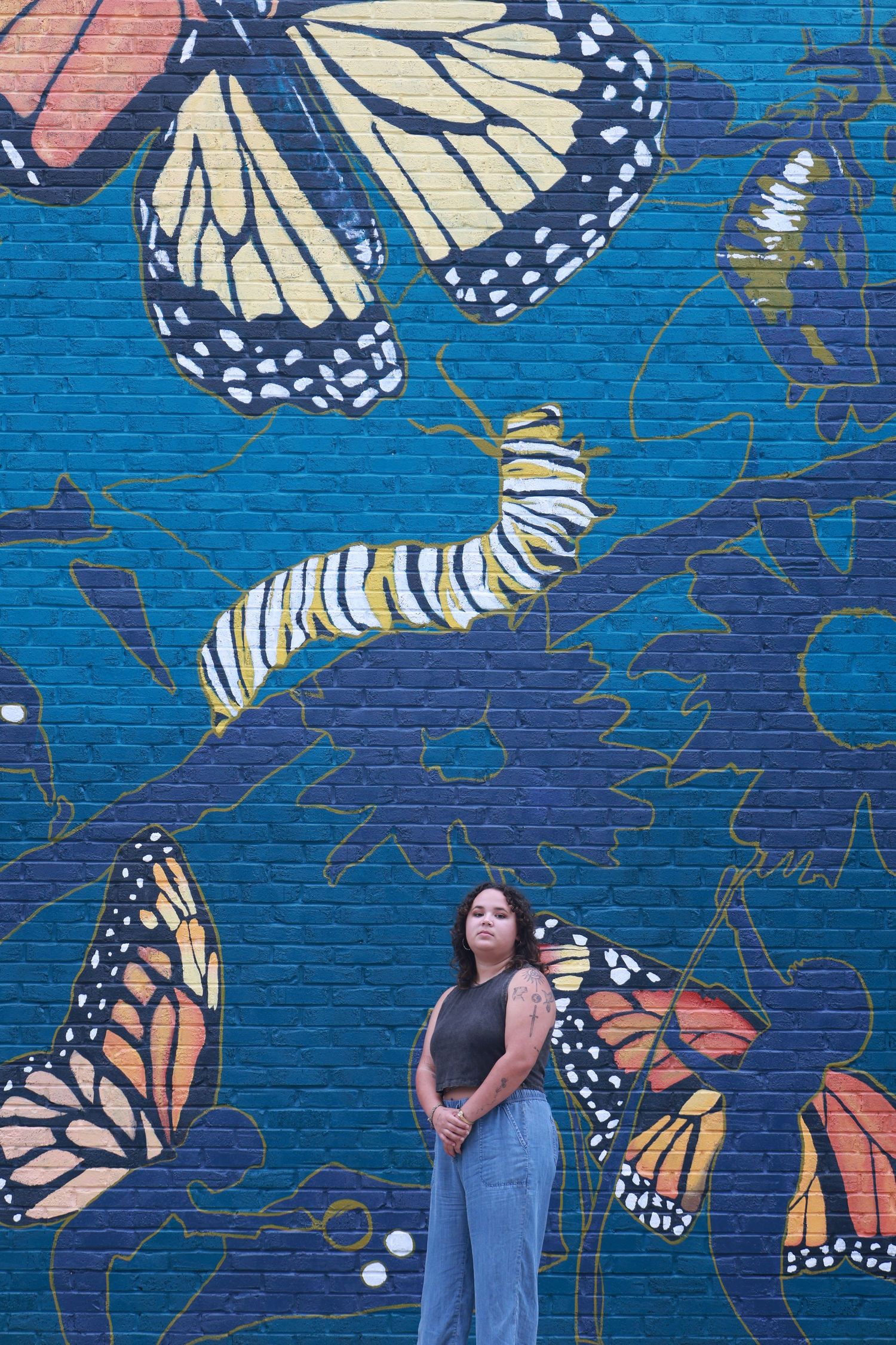 Woman standing before a vibrant blue brick wall mural featuring monarch butterflies and caterpillars in various life stages.