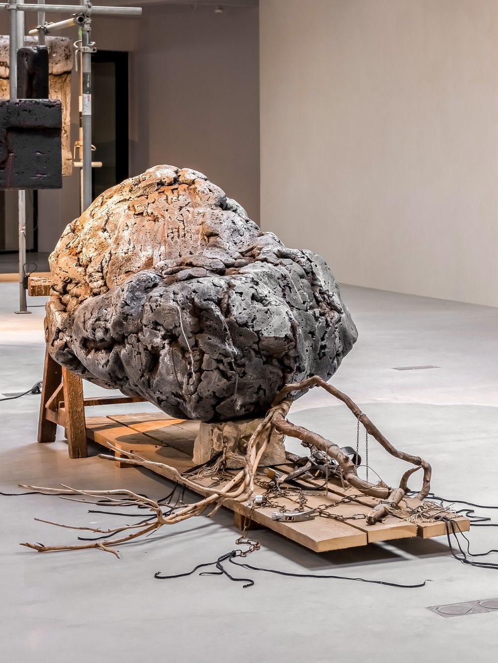 Large rough boulder sculpture on wooden pallet with tangled roots in a contemporary art gallery.