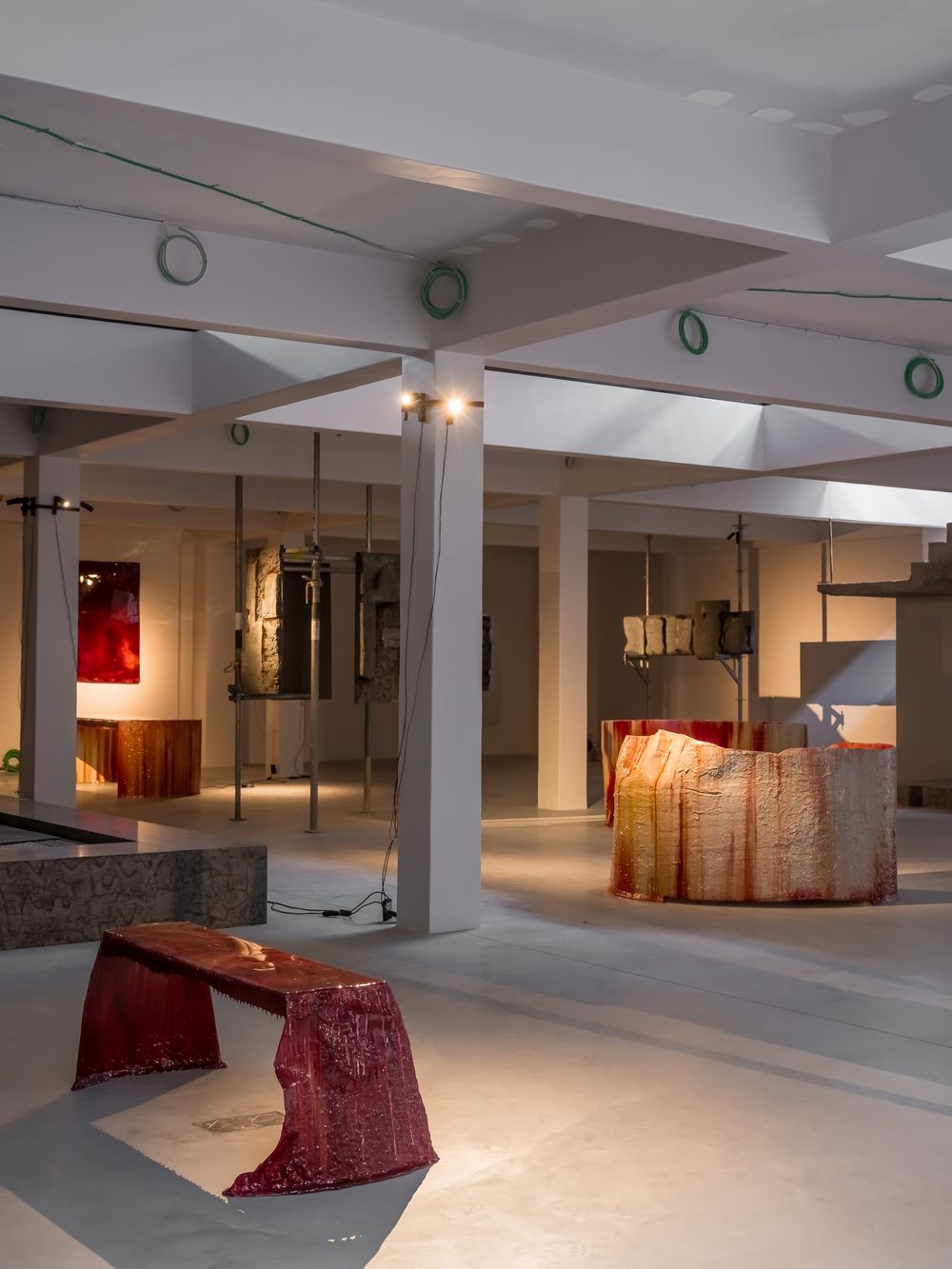 Modern art gallery interior with red sculptural furniture pieces, white columns, and mixed media installations on display.