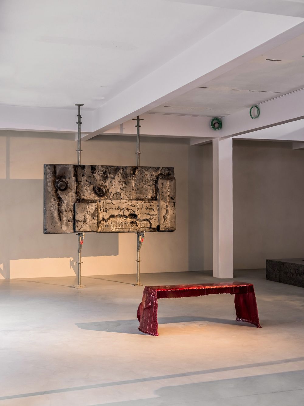 Contemporary art installation featuring a suspended textured panel on metal poles above a red bench in a minimalist gallery space.