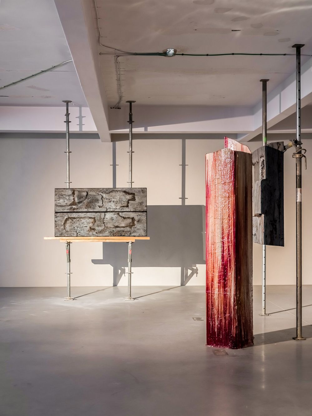 Contemporary art installation featuring sculptural works including a stone relief on a stand and a red textured column in a gallery.