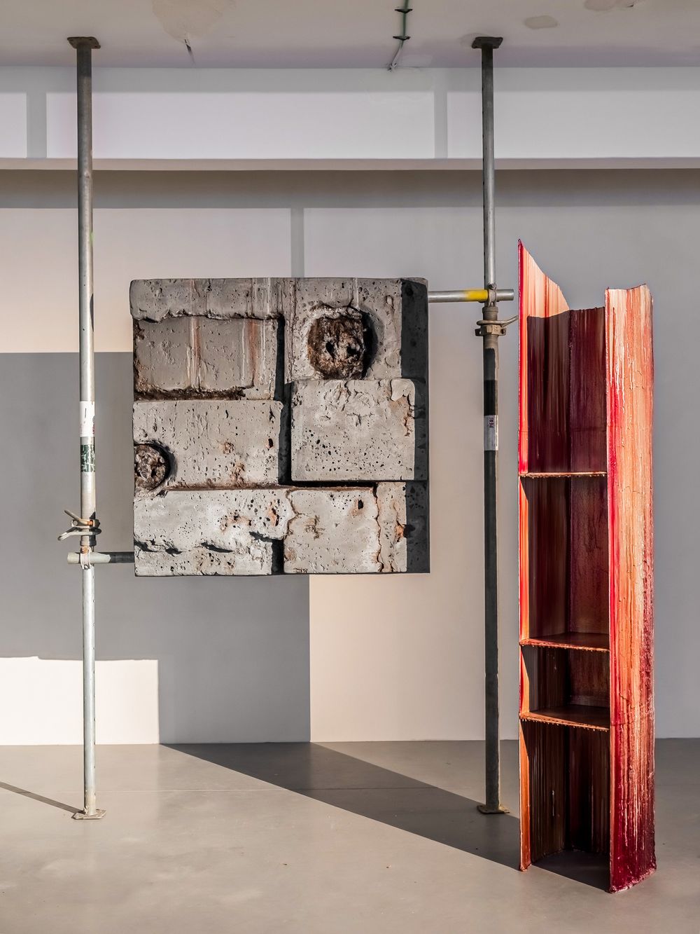Contemporary art installation featuring suspended concrete blocks on metal poles beside a red wooden shelving unit in a gallery.