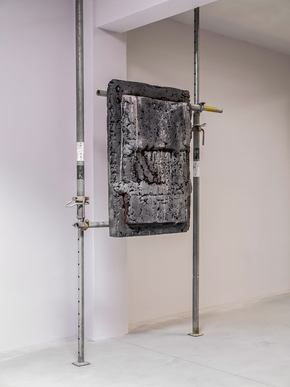 A textured dark sculpture mounted on metal scaffolding poles displayed in a white gallery space.