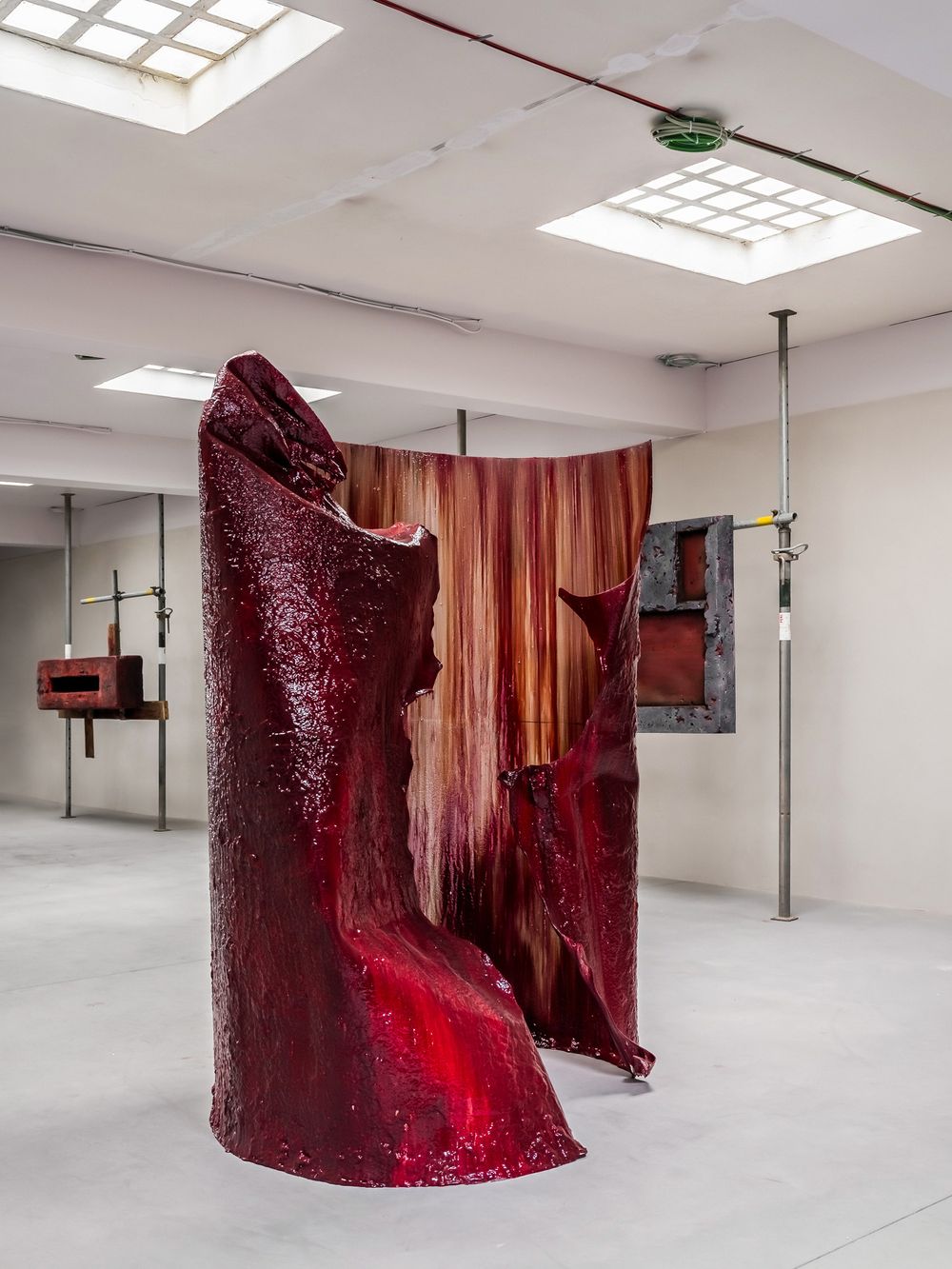 Large red sculptural installation with organic flowing forms displayed in a white gallery space with skylight windows.