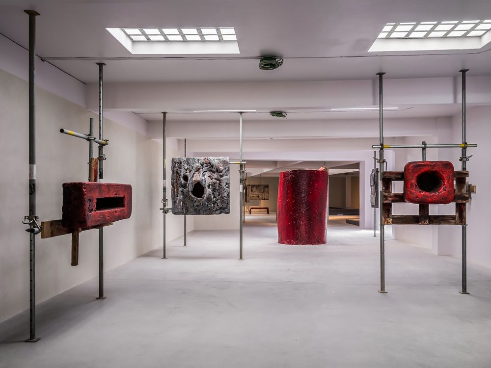 Contemporary art gallery featuring red sculptural installations mounted on metal poles with abstract artwork displayed on white walls.