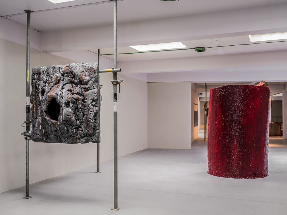 Modern art gallery featuring a textured gray wall sculpture on metal poles and a large red cylindrical artwork.