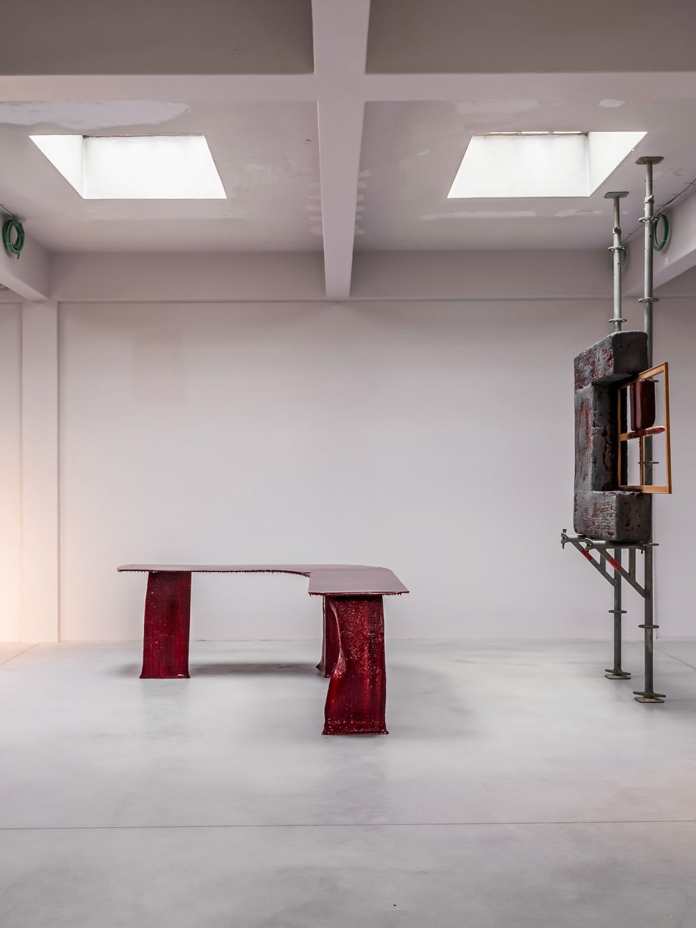 Modern art installation featuring a red sculptural table and industrial metal tower structure in a minimalist white gallery space.