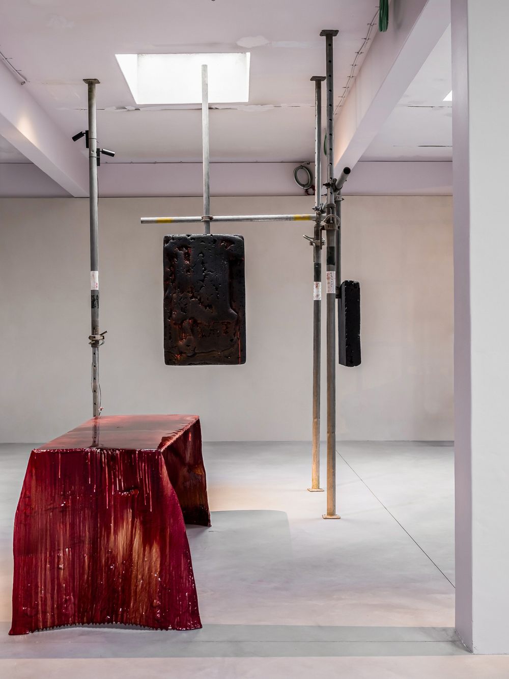 Contemporary art installation with red resin table, dark textured panel, and metal poles in a white gallery space.