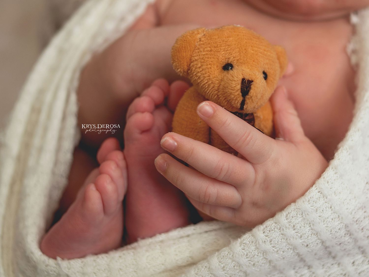 Newborn baby feet and tiny hands holding a small orange knit teddy bear wrapped in a white blanket.