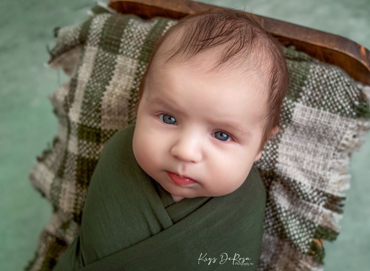 Newborn baby wrapped in green swaddle blanket lying in wooden bowl with plaid blanket backdrop.