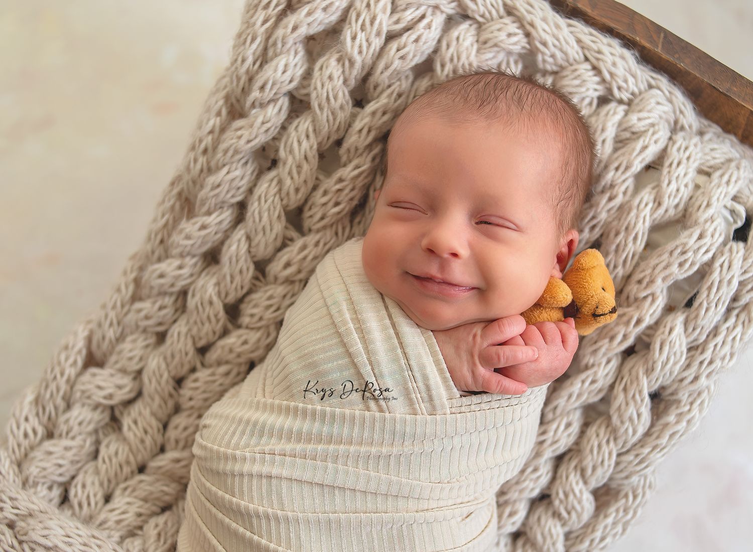 Smiling newborn baby swaddled in cream knit wrap, nestled in chunky knit basket with tiny yellow bear toy.