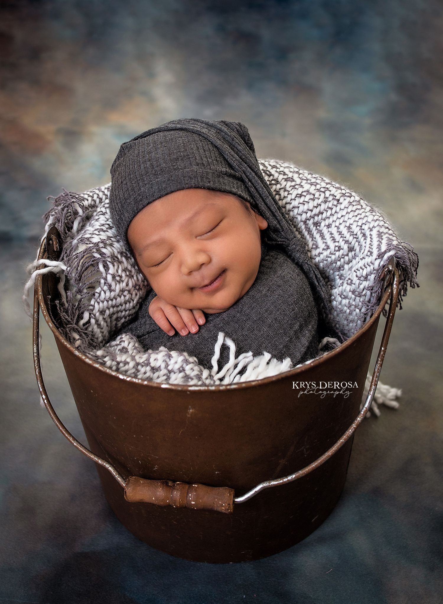 Smiling newborn baby sleeping in a rustic metal bucket wrapped in gray fabric with matching hat.