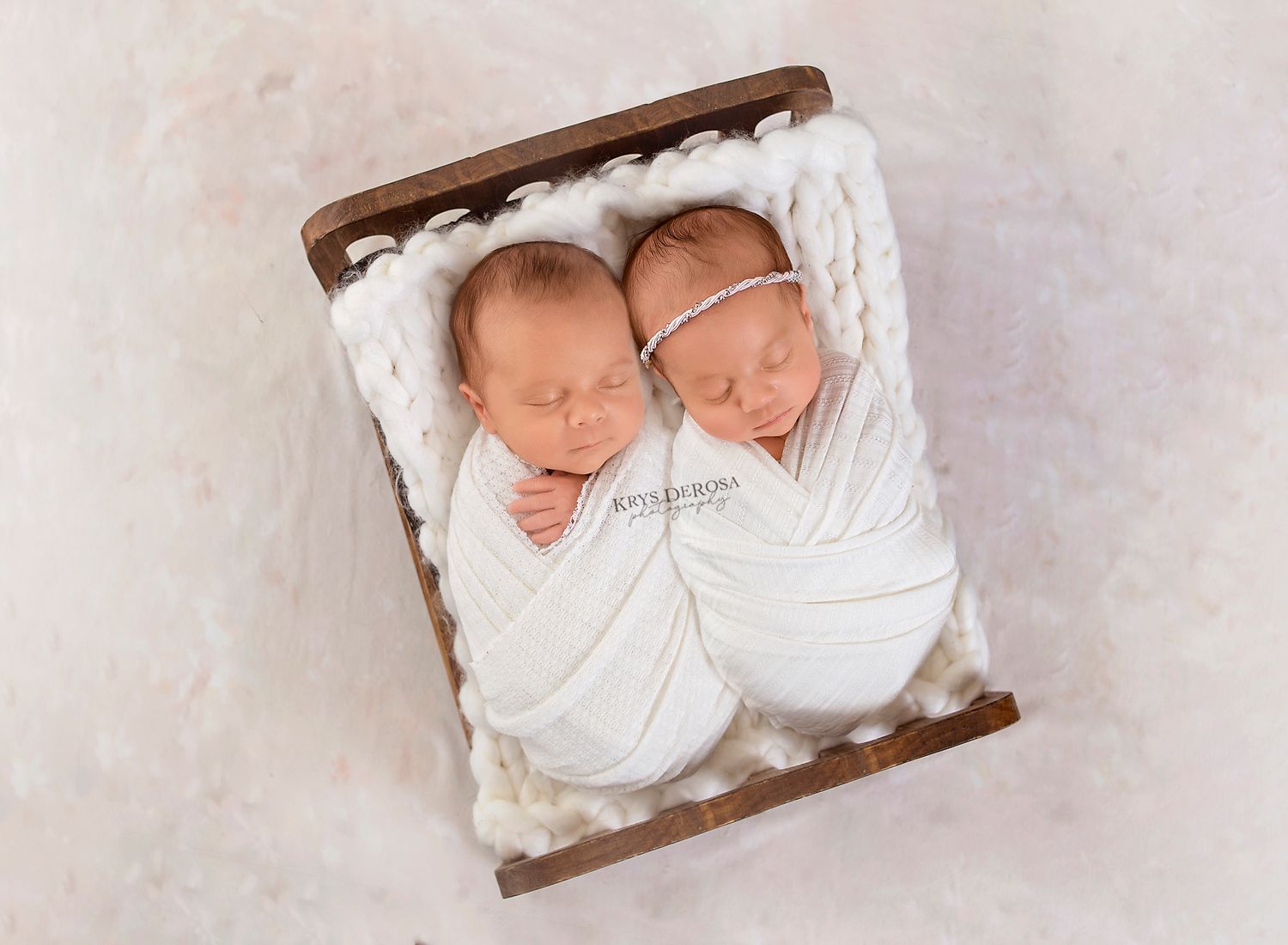 Two newborn twins swaddled in white wraps sleeping peacefully side by side in a small wooden bed.