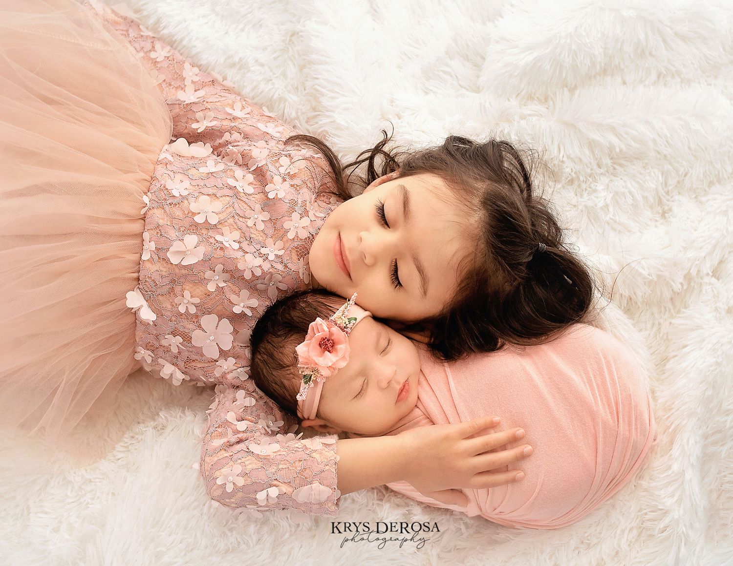 A young girl in a pink floral dress cuddles a sleeping newborn baby wrapped in pink on a white fur blanket.