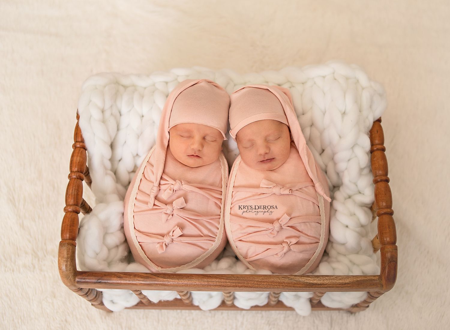Two newborn twins swaddled in pink wraps and hats sleep peacefully side by side in a wooden basket.