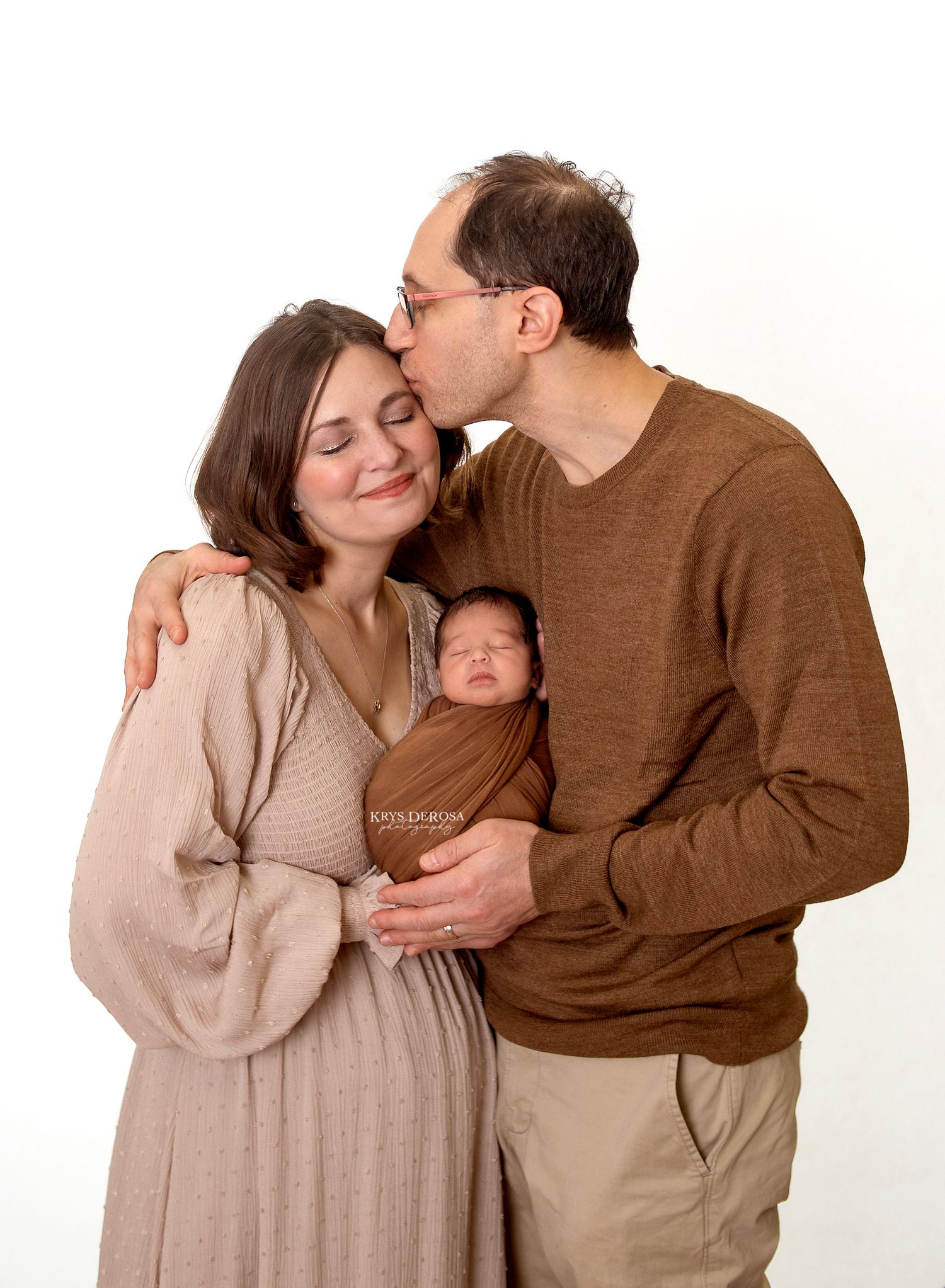 Parents posing with newborn baby as father kisses mother on cheek against white background.