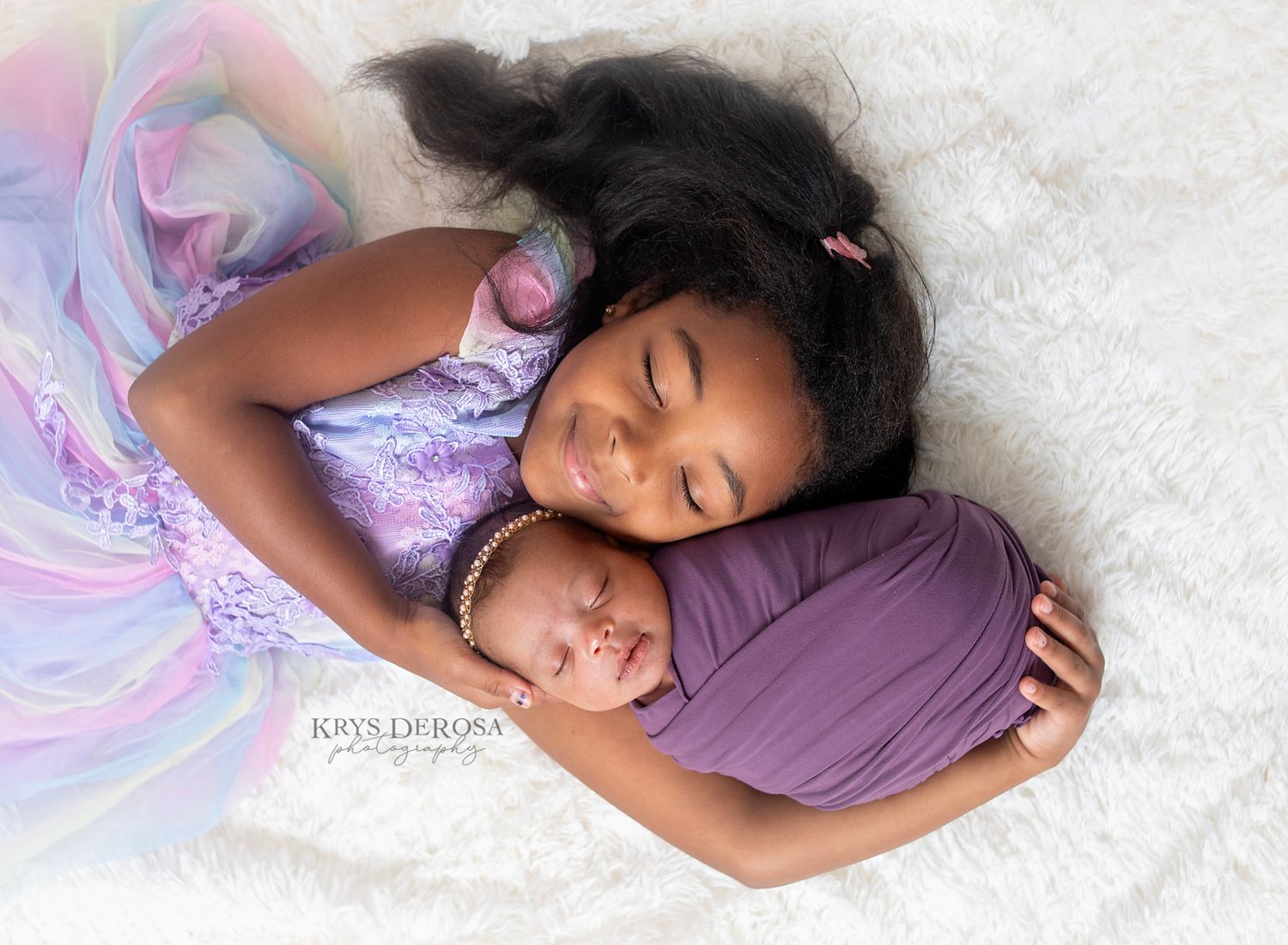Young girl lying down smiling with eyes closed while holding swaddled newborn baby in purple wrap.