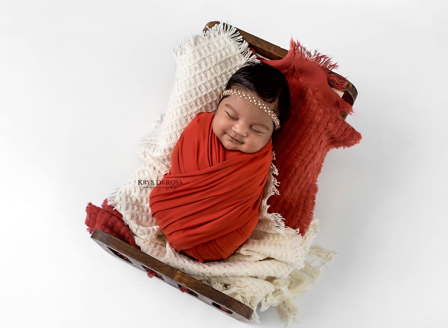Smiling newborn baby wrapped in red swaddle cloth resting in a rustic wooden crate.