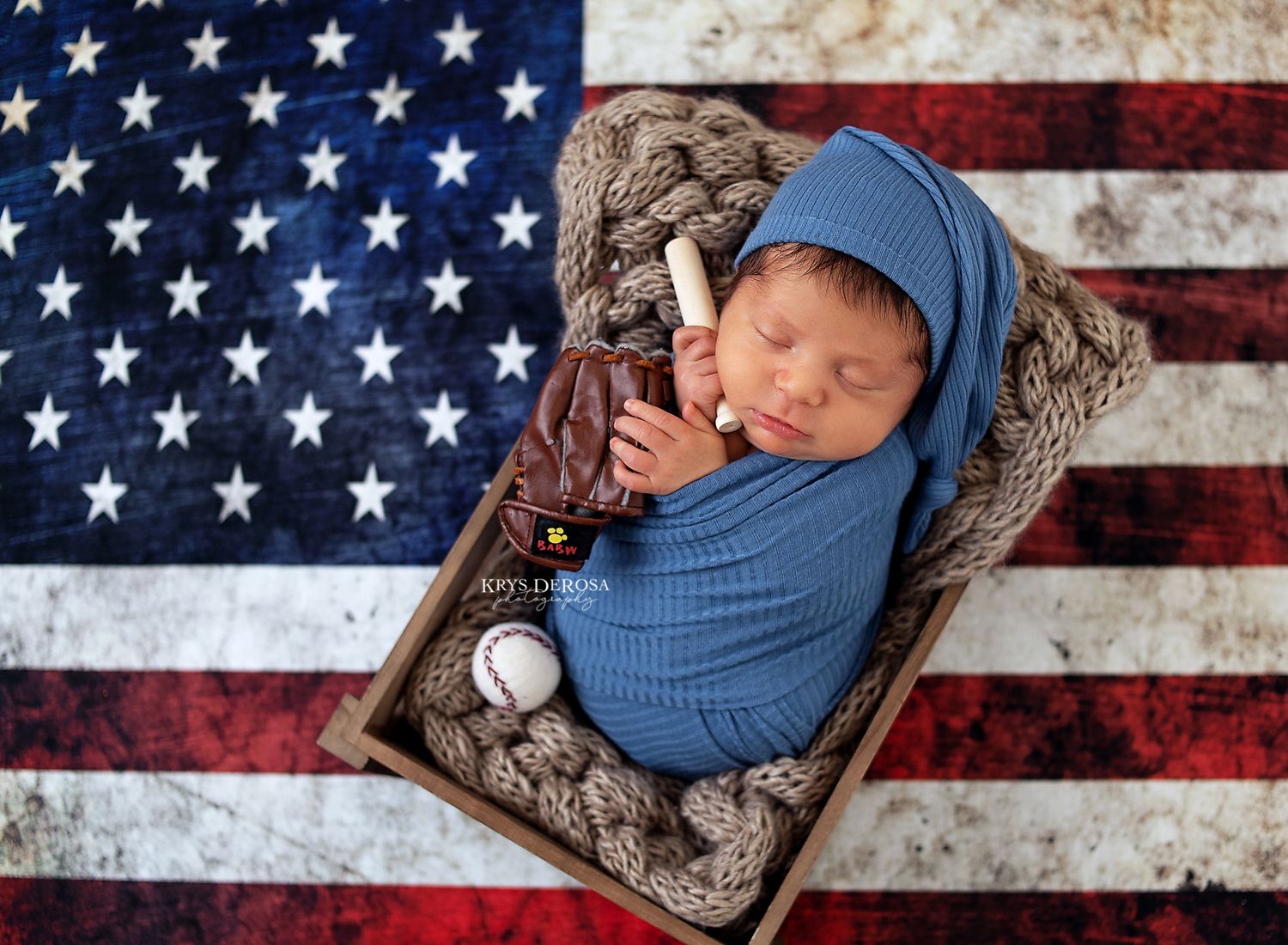 Sleeping newborn wrapped in blue swaddle posed with baseball glove and bat on American flag backdrop.