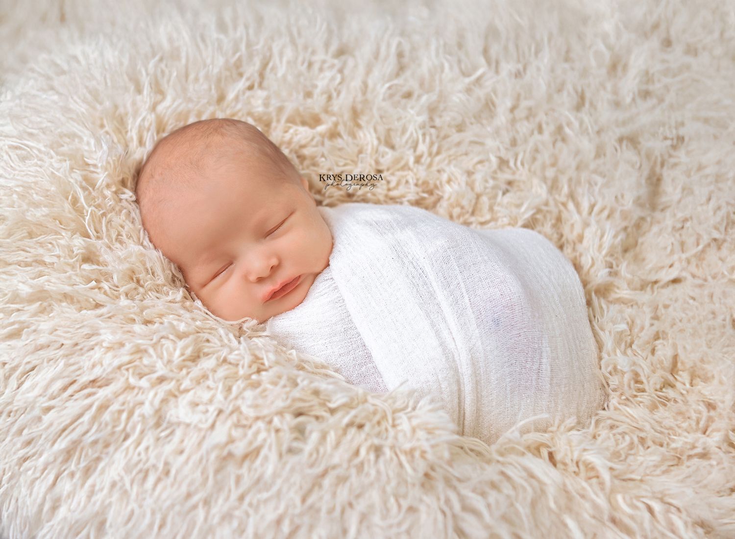 Peaceful newborn baby wrapped in white swaddle sleeping on a fluffy cream-colored fur blanket.