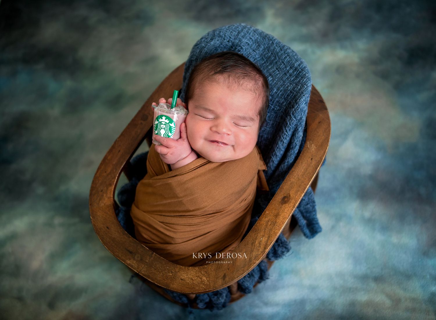 Smiling newborn wrapped in brown swaddle wearing blue knit bonnet, holding tiny Starbucks cup in wooden bowl prop.