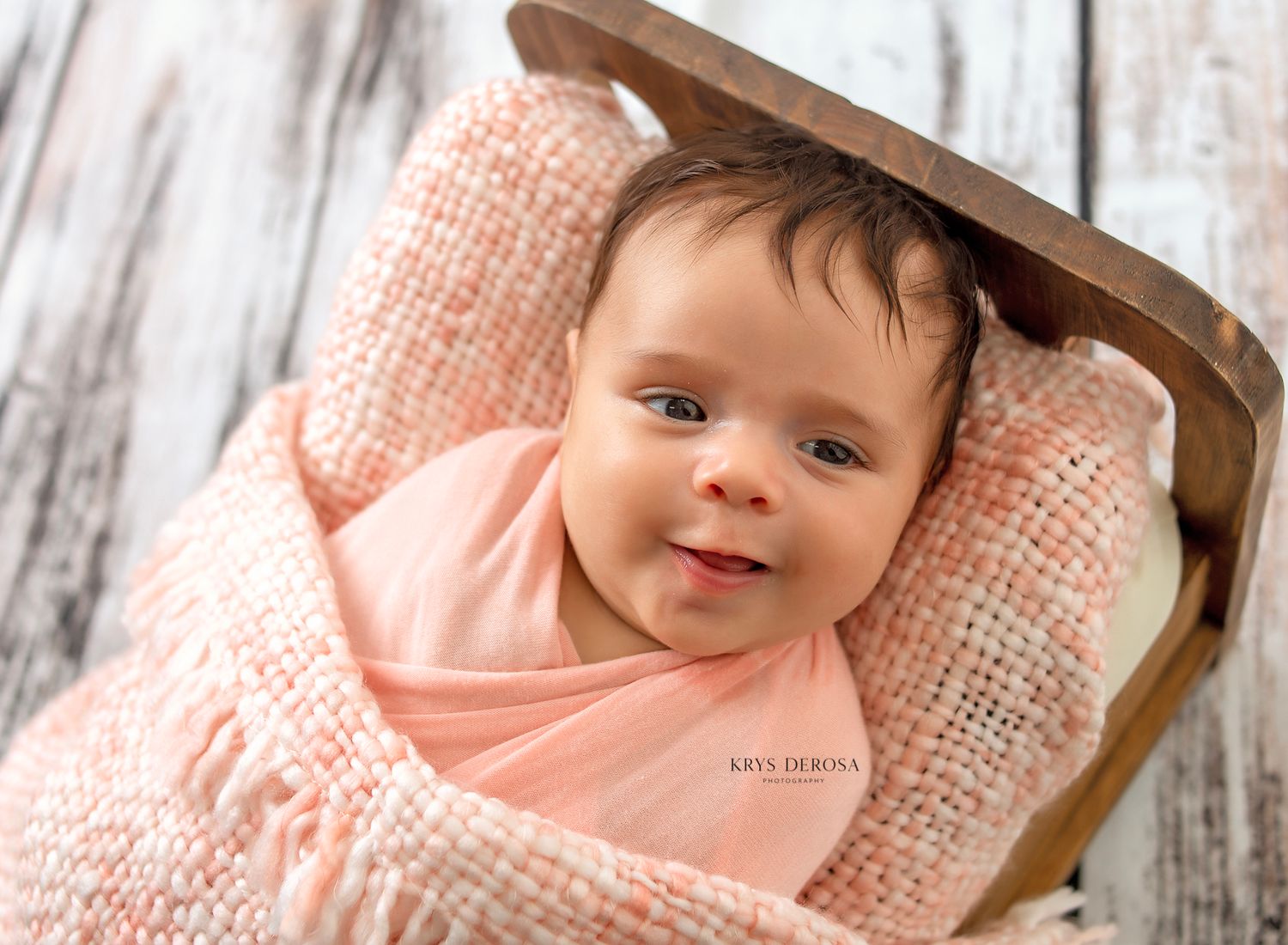 Newborn baby wrapped in soft pink knit blanket, smiling in a wooden bed on a rustic white background.