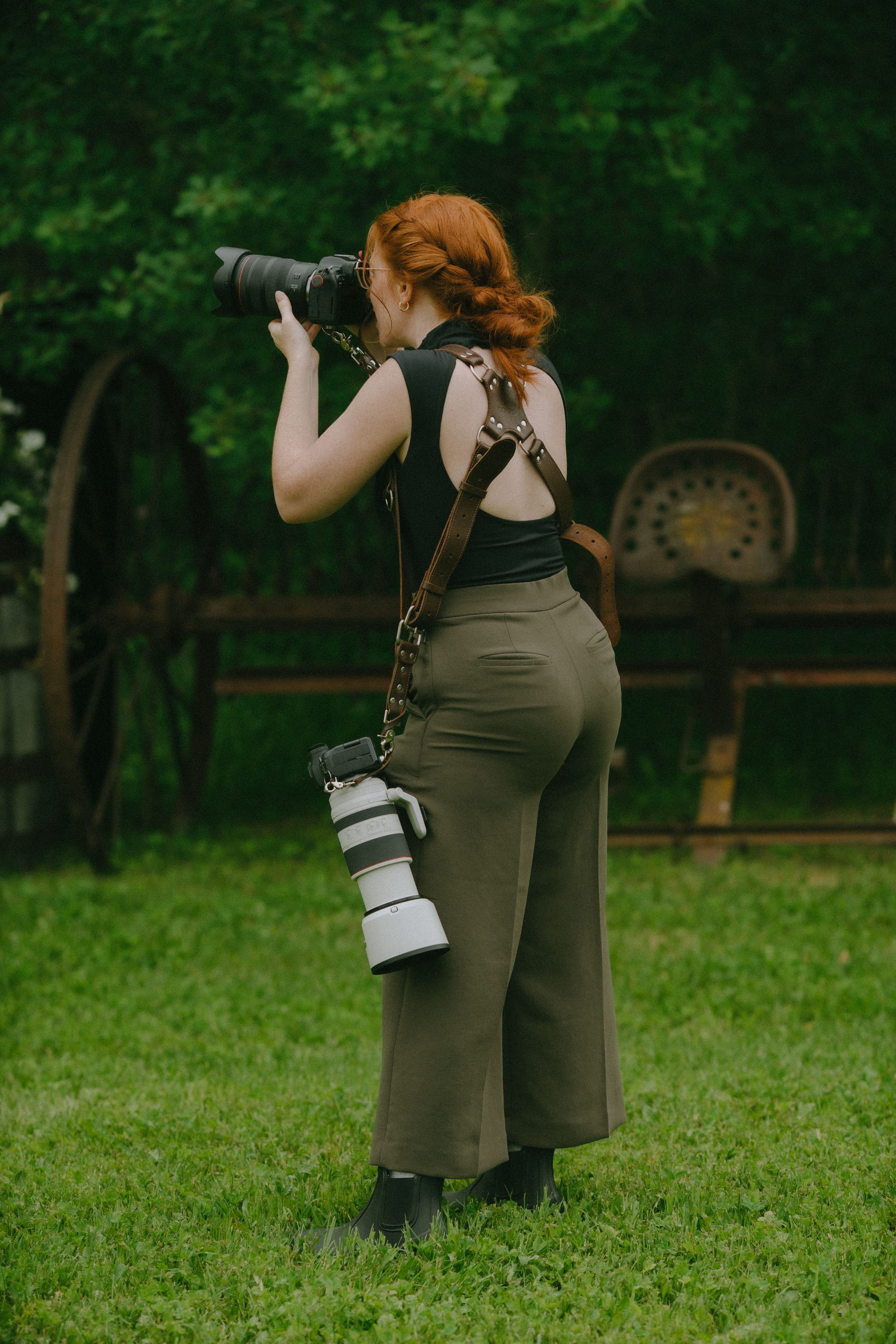 Red-haired female photographer shooting with DSLR camera outdoors, second lens hanging from strap, standing on green grass.