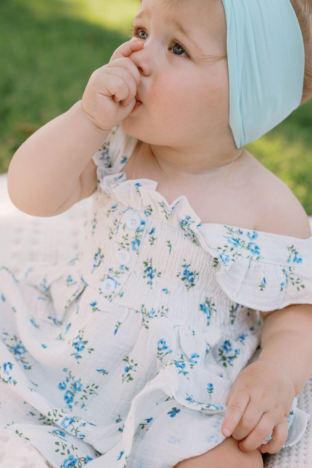 Adorable baby girl in floral off-shoulder dress with blue headband sitting outdoors.