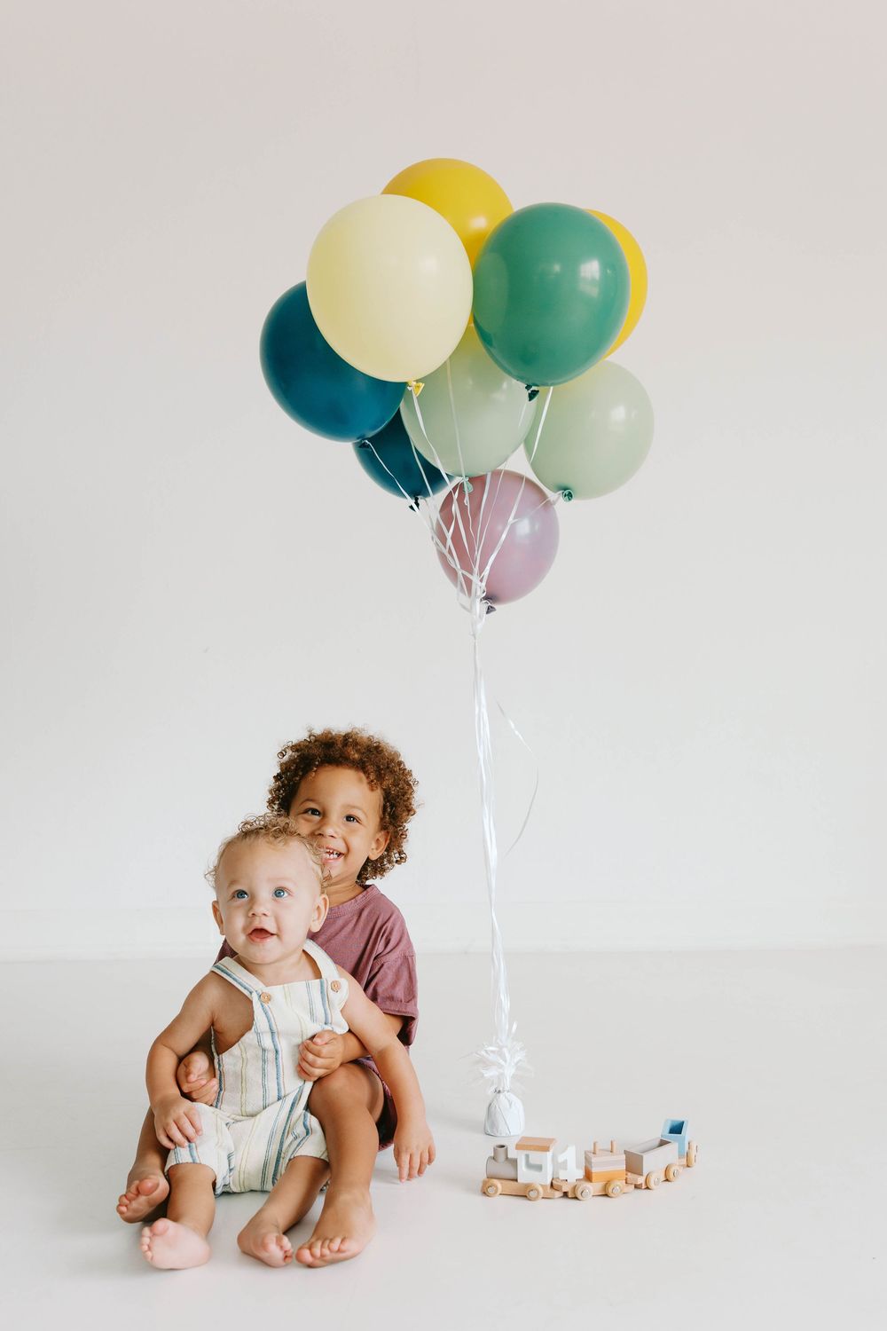 Two young children sit together with colorful balloons and a toy train on a white background.