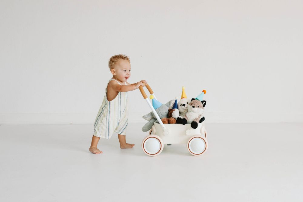 Toddler in a striped romper pushing a white wooden wagon filled with stuffed animals.