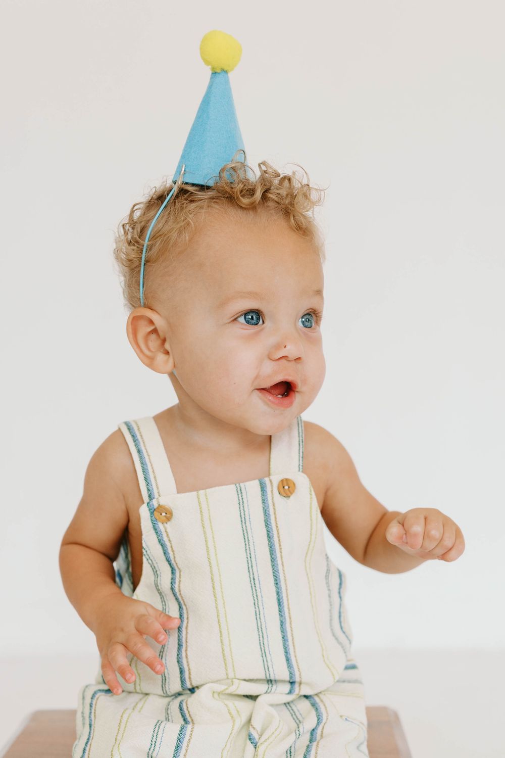 Curly-haired toddler in a striped romper wearing a blue party hat with a yellow pompom.