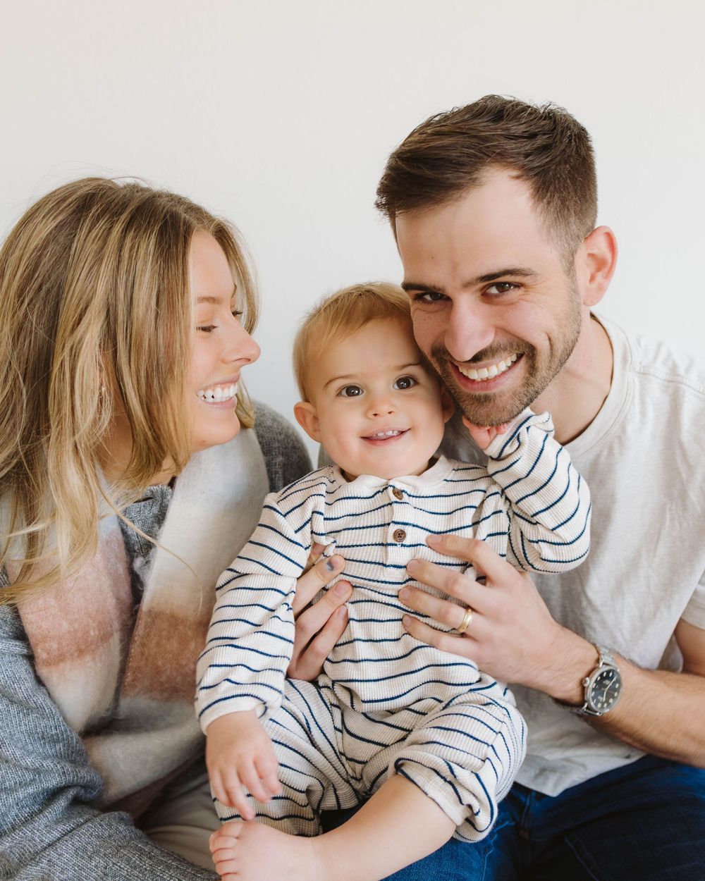 Smiling young family with a toddler in a striped outfit posing together against a white background.