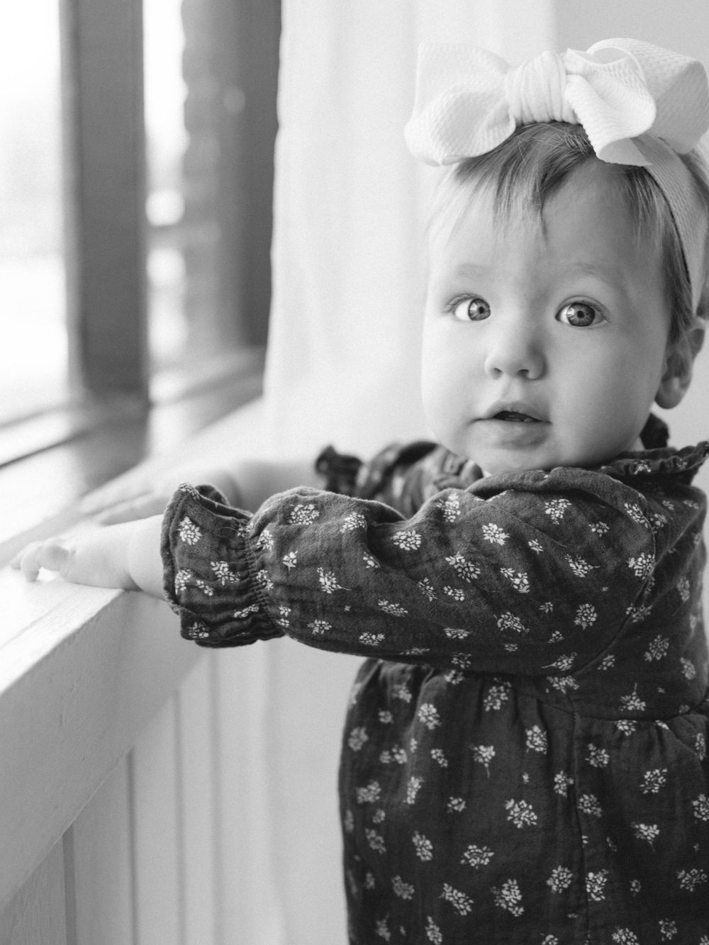 Black and white photo of a baby girl in a floral dress and white bow headband standing at a window.